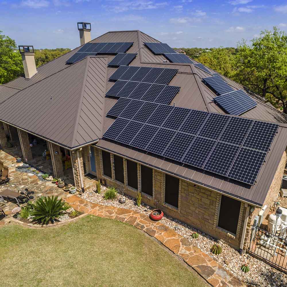 Aerial view of a house with a brown metal roof covered in solar panels under a partly cloudy blue sky.