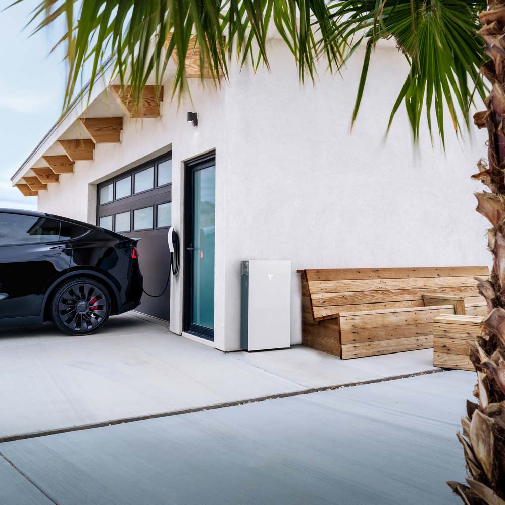 Black Tesla charging at a wall-mounted station beside a modern house with wooden benches, a palm tree, and a Tesla Powerwall 3.