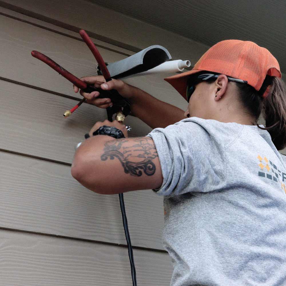 Freedom Power HVAC Installer wearing an orange cap and sunglasses installing HVAC pipes on a house exterior.