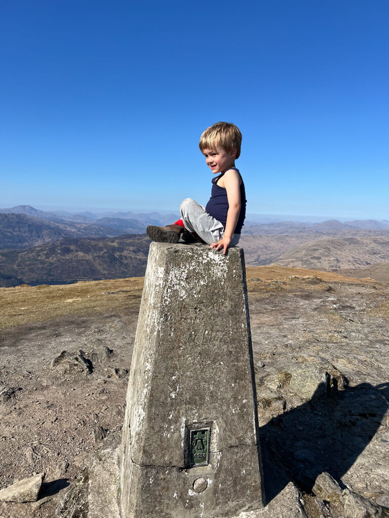 A young boy in a navy tank top and gray pants sitting atop a weathered trig point on a mountain under a clear blue sky.