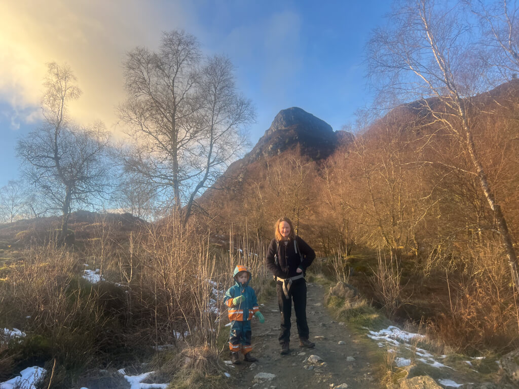 Woman and child standing on a rocky trail surrounded by bare trees and patches of snow with a mountain in the background under a partly cloudy sky.