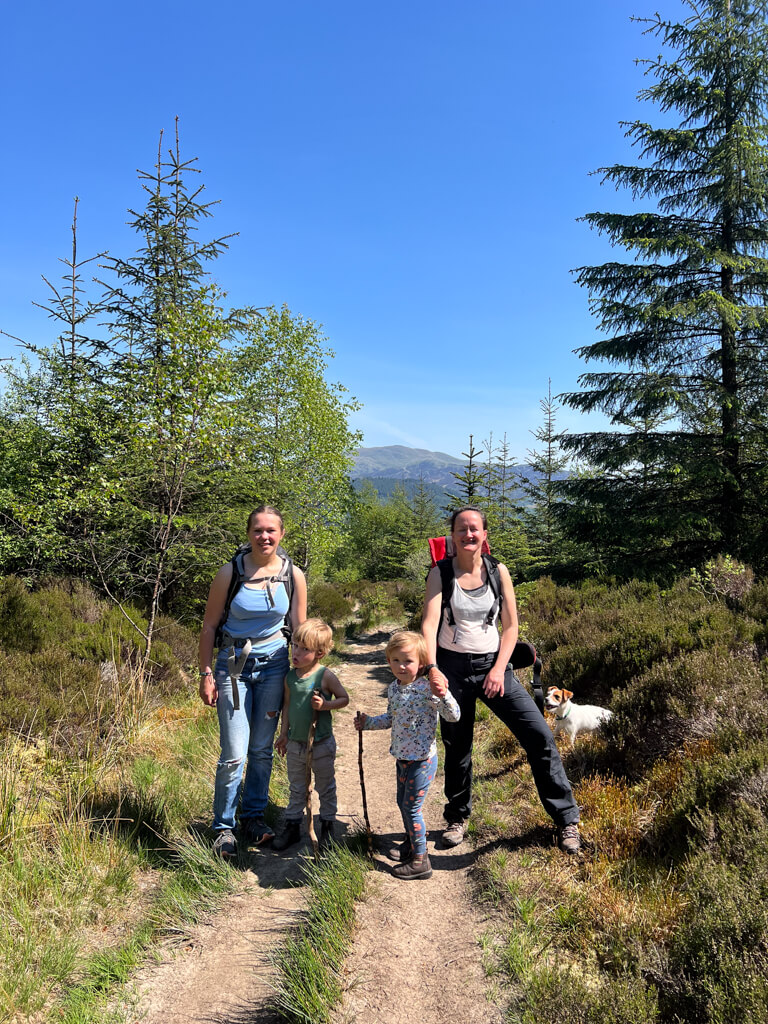 Two women and two children standing on a forest trail surrounded by green trees and shrubs under a clear blue sky, with a small dog nearby.