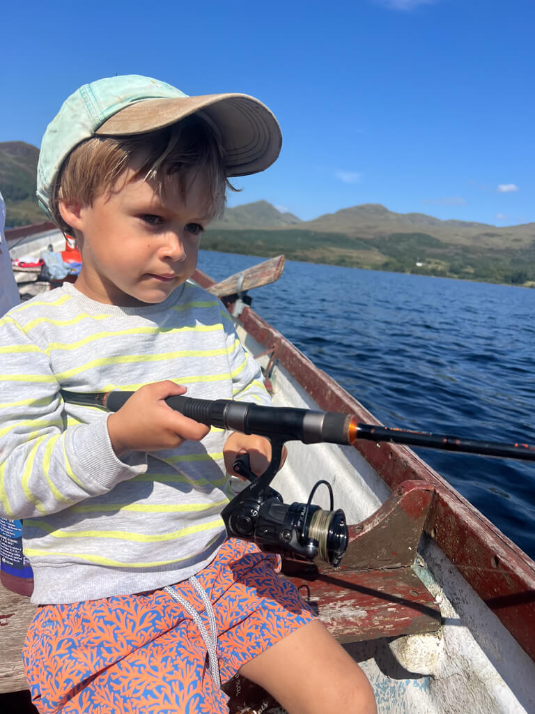 Young boy wearing a cap and striped sweater holding a fishing rod while sitting in a boat on a lake with mountains in the background.