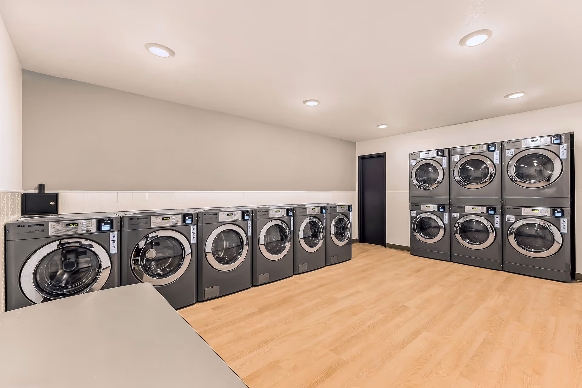 Modern laundry room with two rows of front-loading washing machines and dryers, wood flooring, white walls, and recessed ceiling lights.