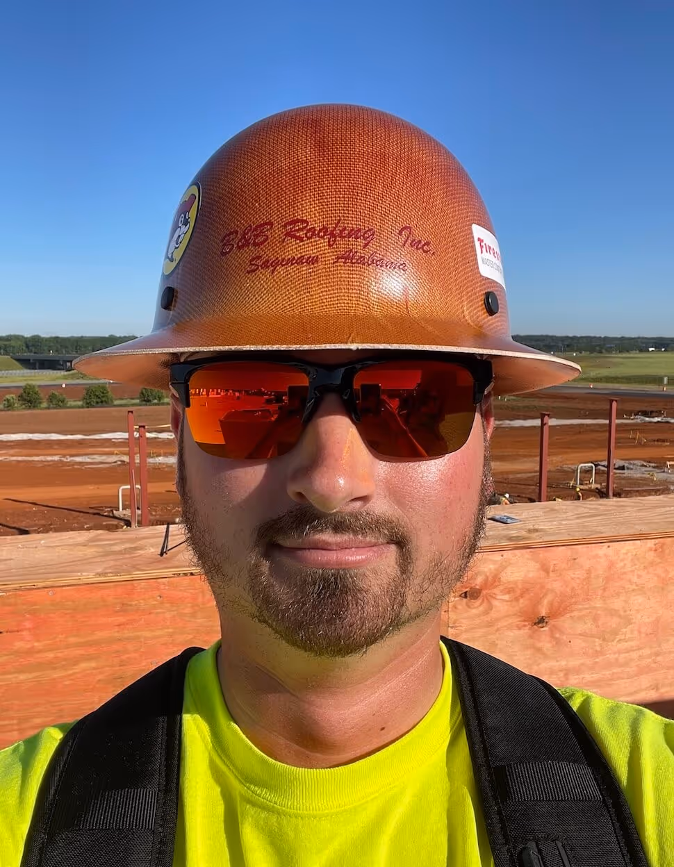 Construction worker wearing an orange hard hat, reflective sunglasses, and a bright yellow shirt at a work site under a clear blue sky.