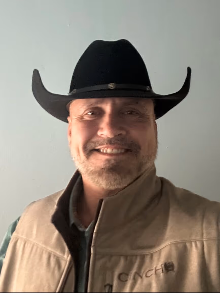 Smiling man wearing a black cowboy hat and beige jacket against a plain light-colored background.