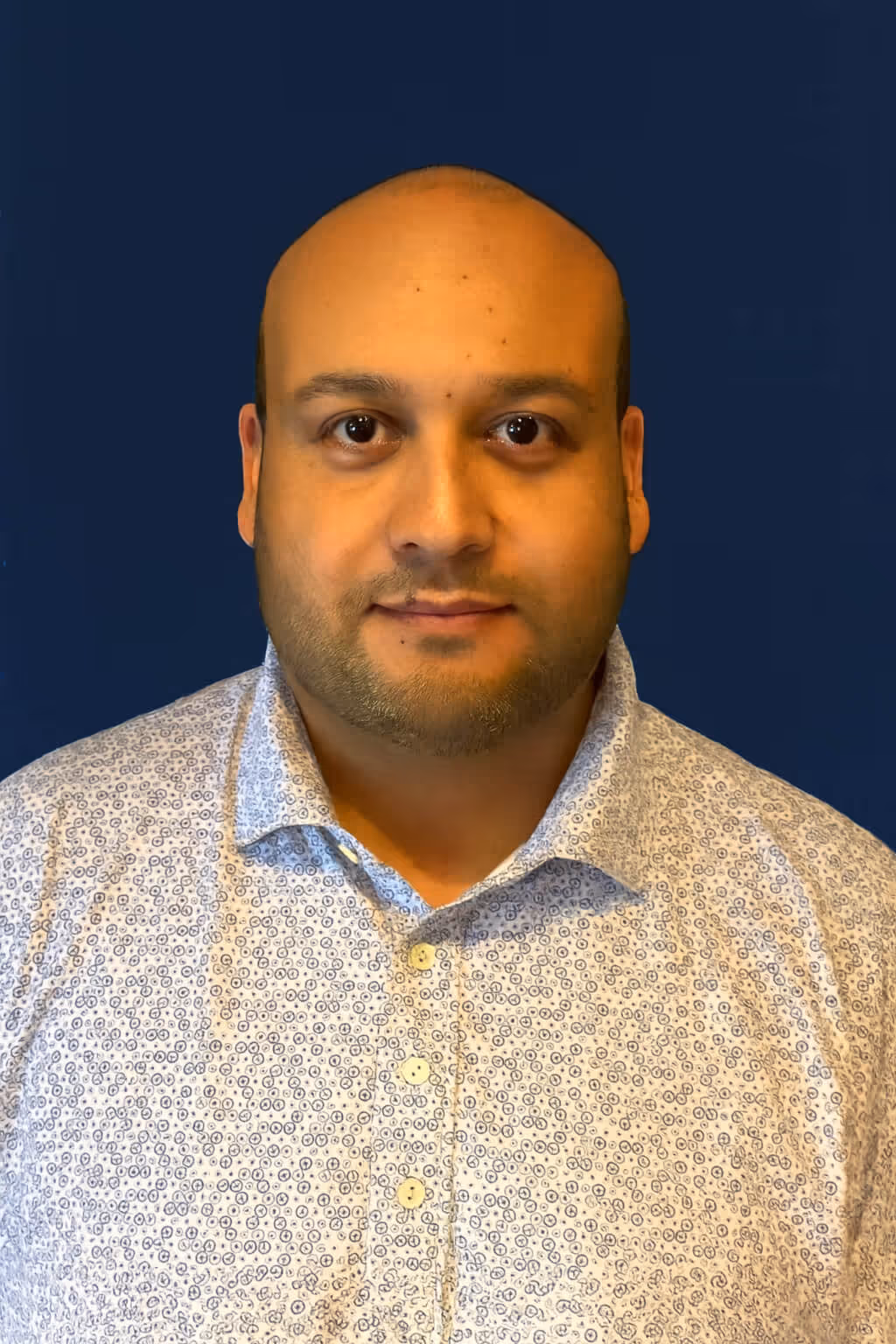 Portrait of a bald man with a slight beard wearing a light patterned button-up shirt against a dark blue background.