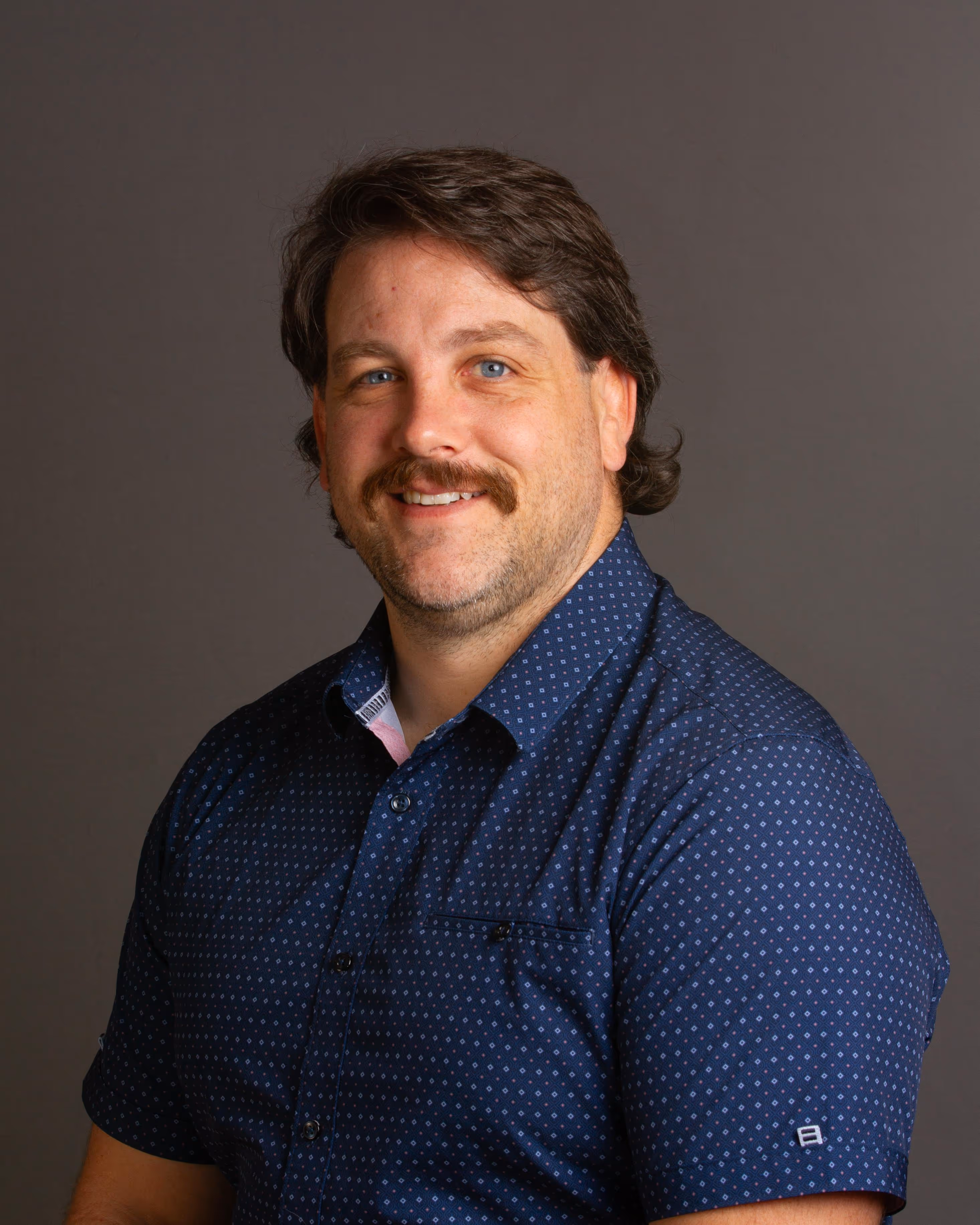 Smiling man with brown hair and mustache wearing a blue patterned shirt against a gray background.