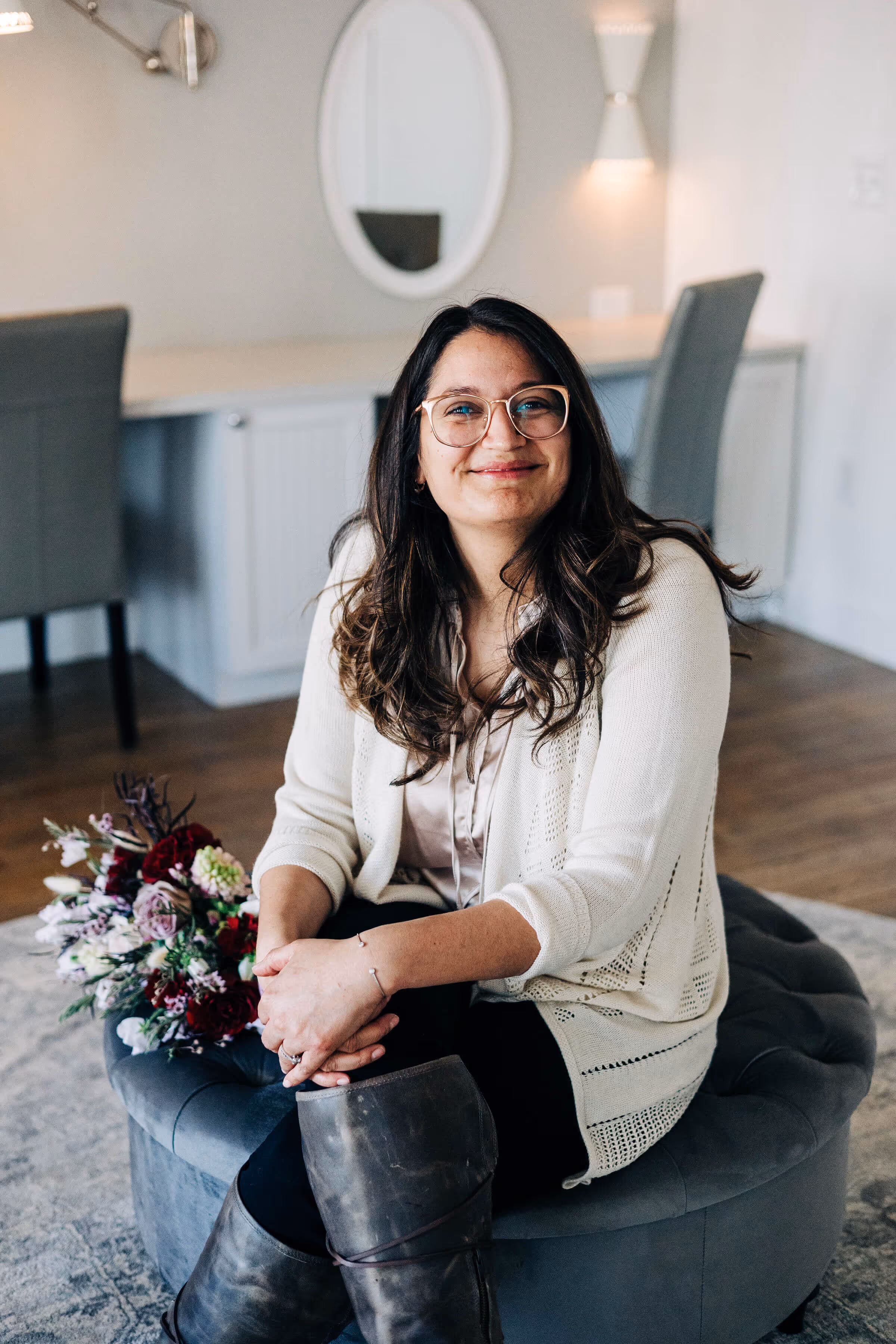 Smiling woman with glasses wearing a white cardigan and boots seated on a gray round ottoman next to a bouquet of flowers in a modern room.