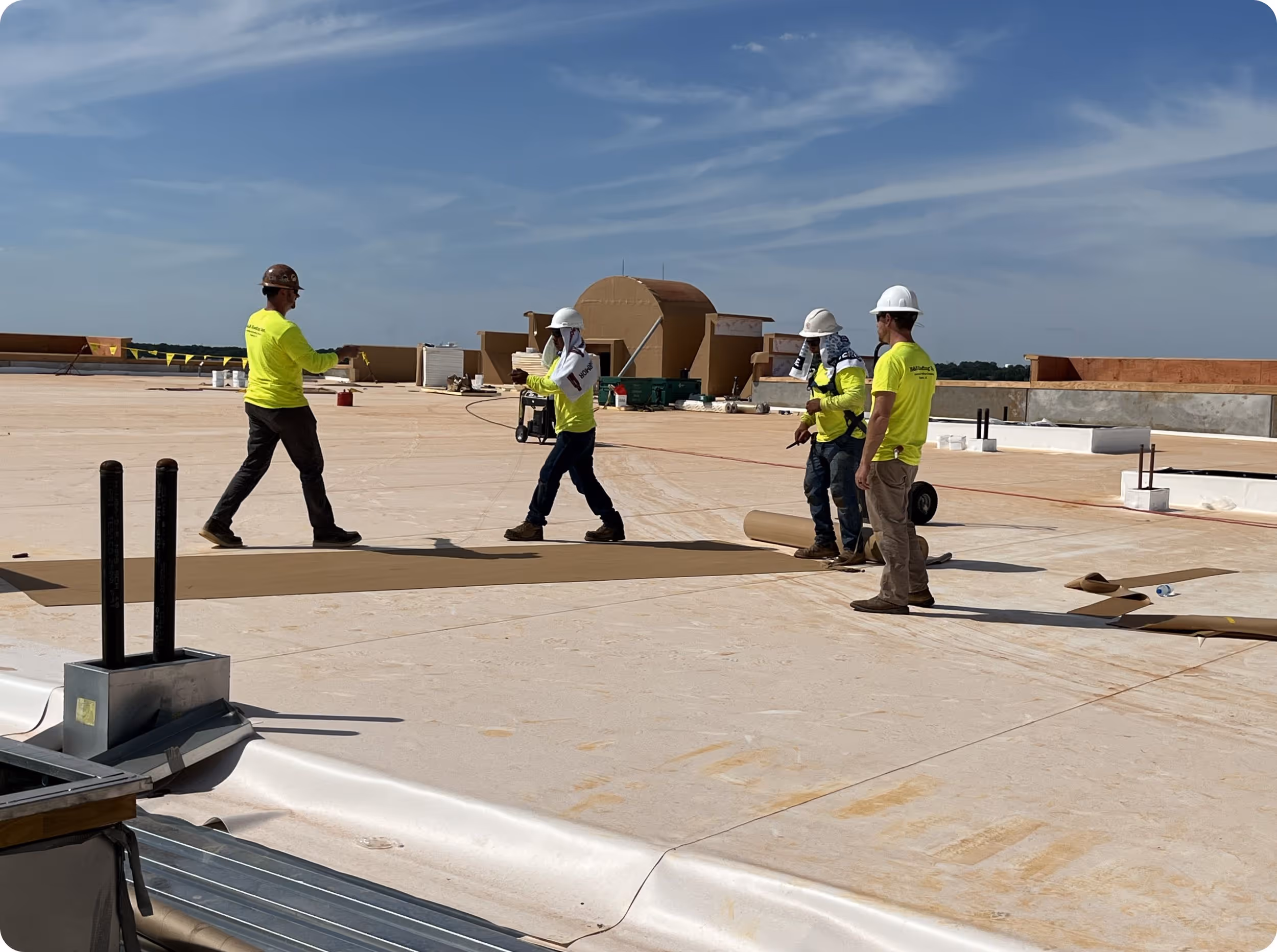 Four construction workers wearing hard hats and bright yellow shirts working on a commercial building rooftop under a clear sky.