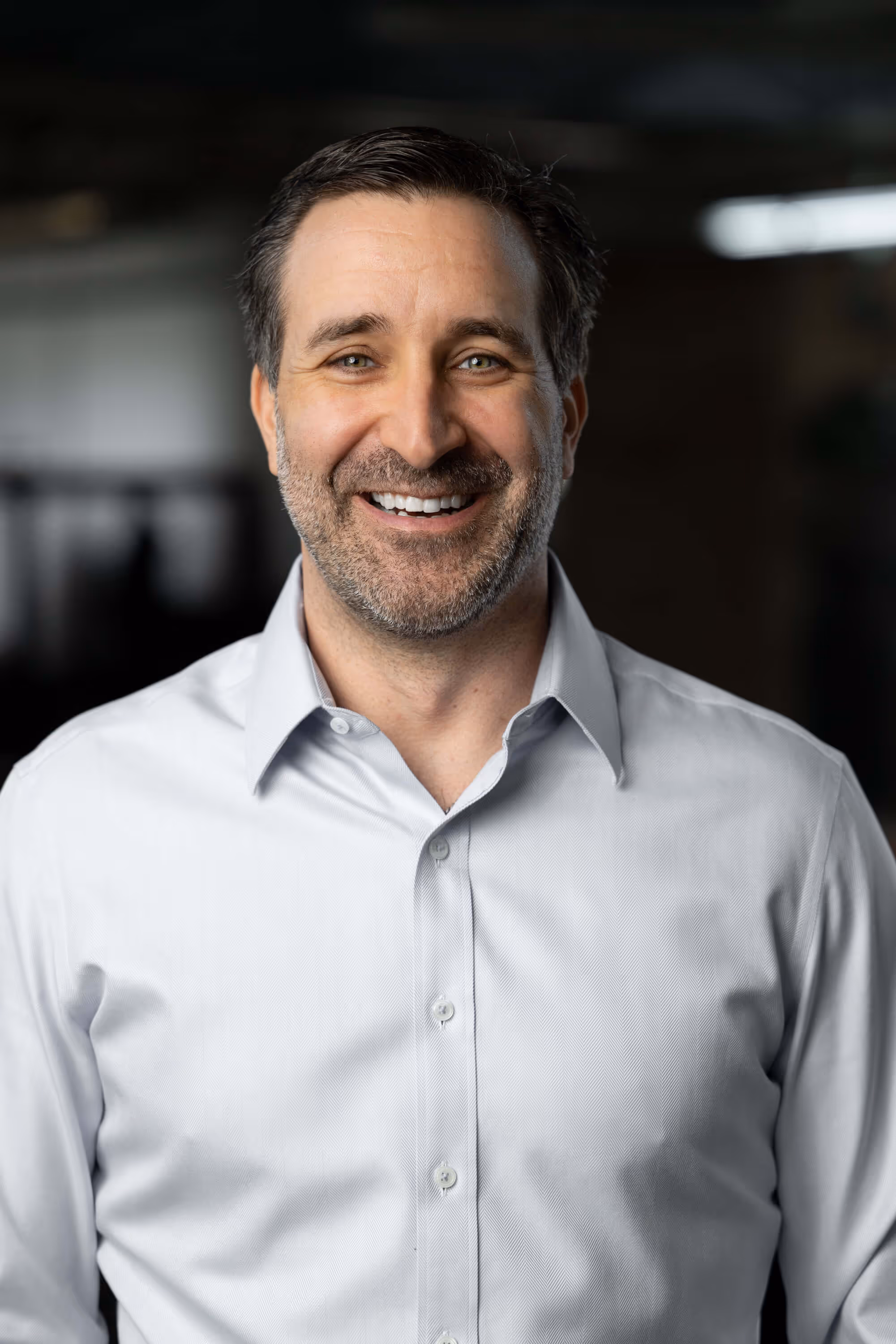 Smiling man with short dark hair and beard wearing a light blue button-up shirt against a dark blurred background.