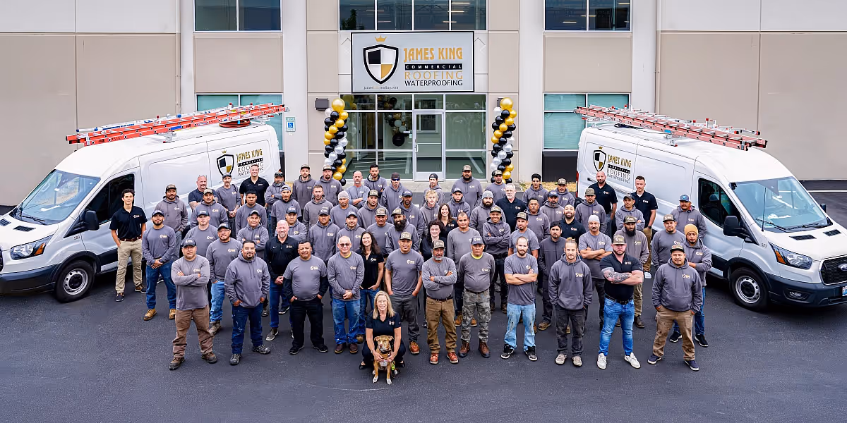 Large group of roofing company employees standing in front of their building with two white vans bearing company logos and ladders on top.
