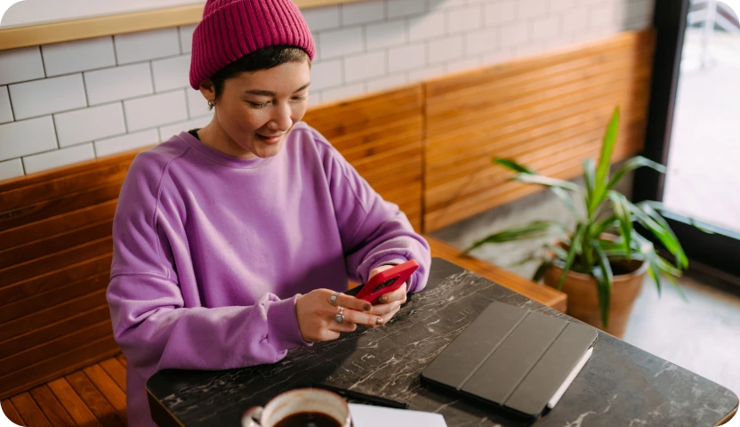 Person in a pink beanie and purple sweatshirt sitting at a table, smiling while using a red smartphone, with a tablet and a coffee cup on the table.