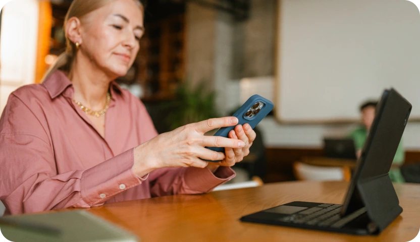 Woman in a pink shirt holding a smartphone while sitting at a table with a tablet in a case and keyboard.
