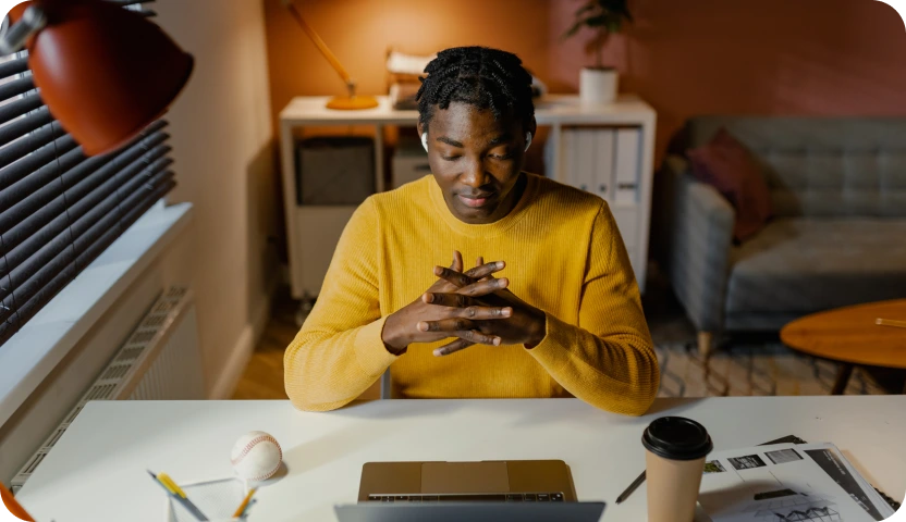 Man in a yellow sweater sitting thoughtfully at a desk with a laptop, coffee cup, baseball, and papers.