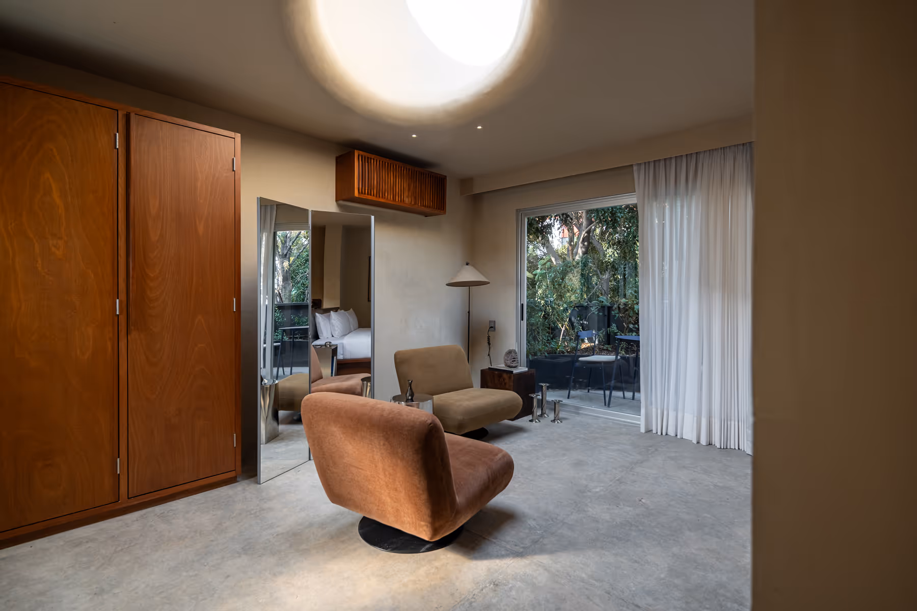 Minimalist living space with two modern chairs, a floor lamp, large mirror, wooden closet, and sliding glass door opening to a patio with greenery.