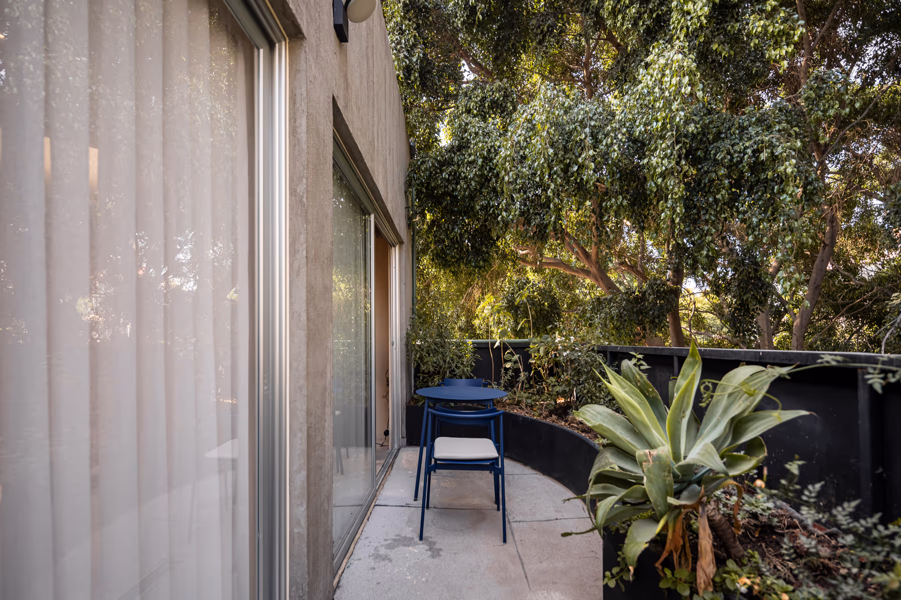 Small outdoor terrace with a blue table and chair set beside sliding glass doors, surrounded by lush green plants and trees.
