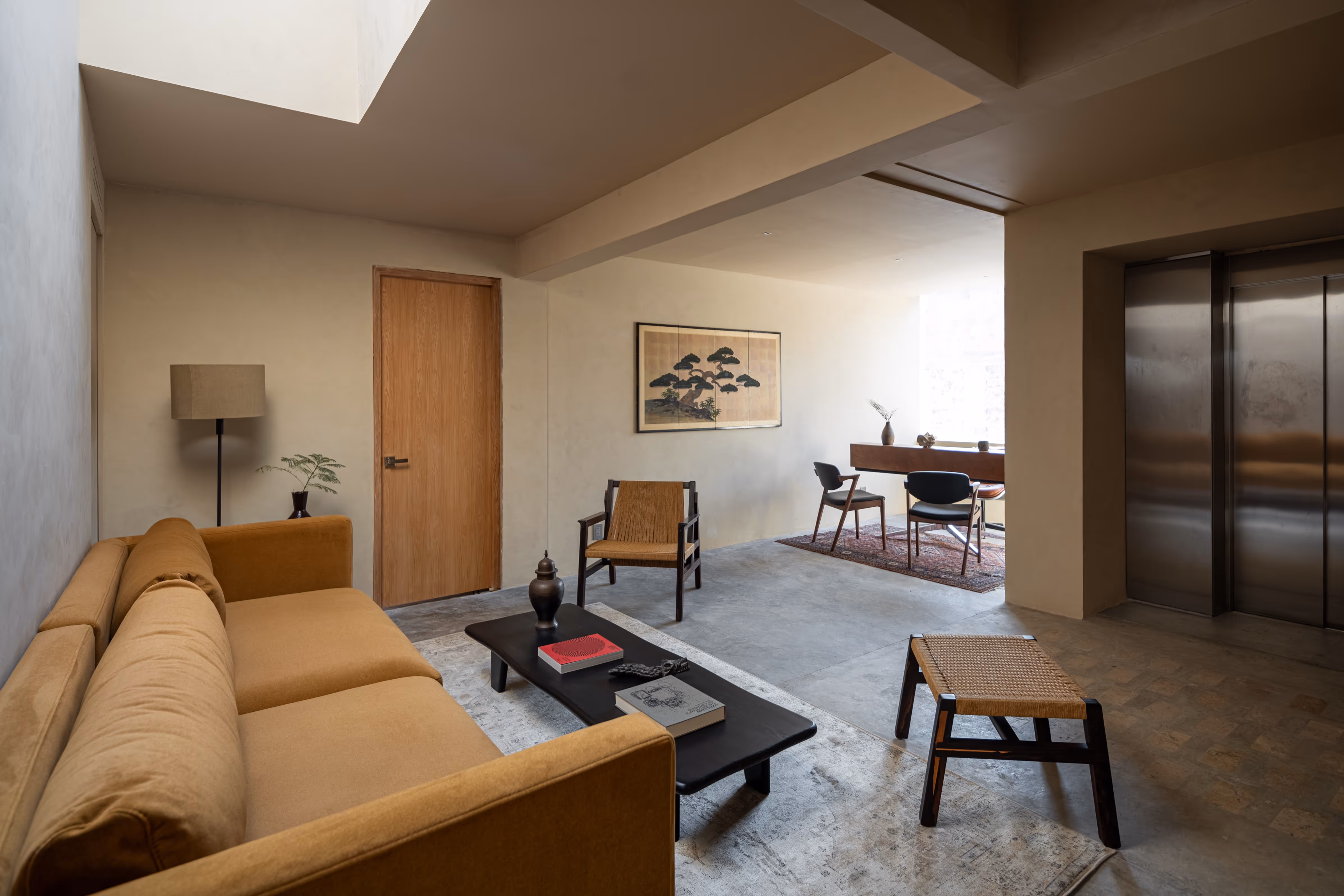 Minimalist reception area with mustard sofa, wooden chair, low black table with books, and elevator doors.