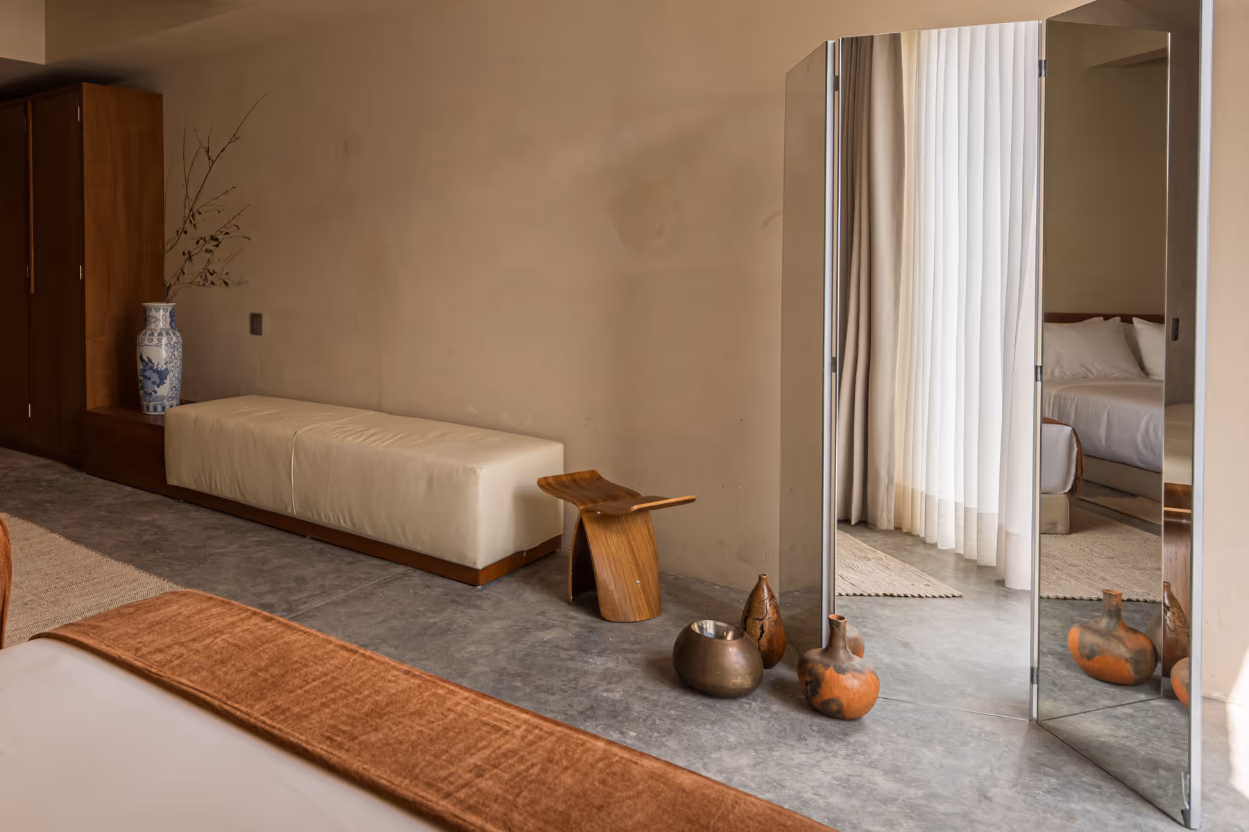 Minimalist bedroom corner with beige bench, wooden stool, decorative vases, and a tall three-panel mirror reflecting a bed and sheer curtains.
