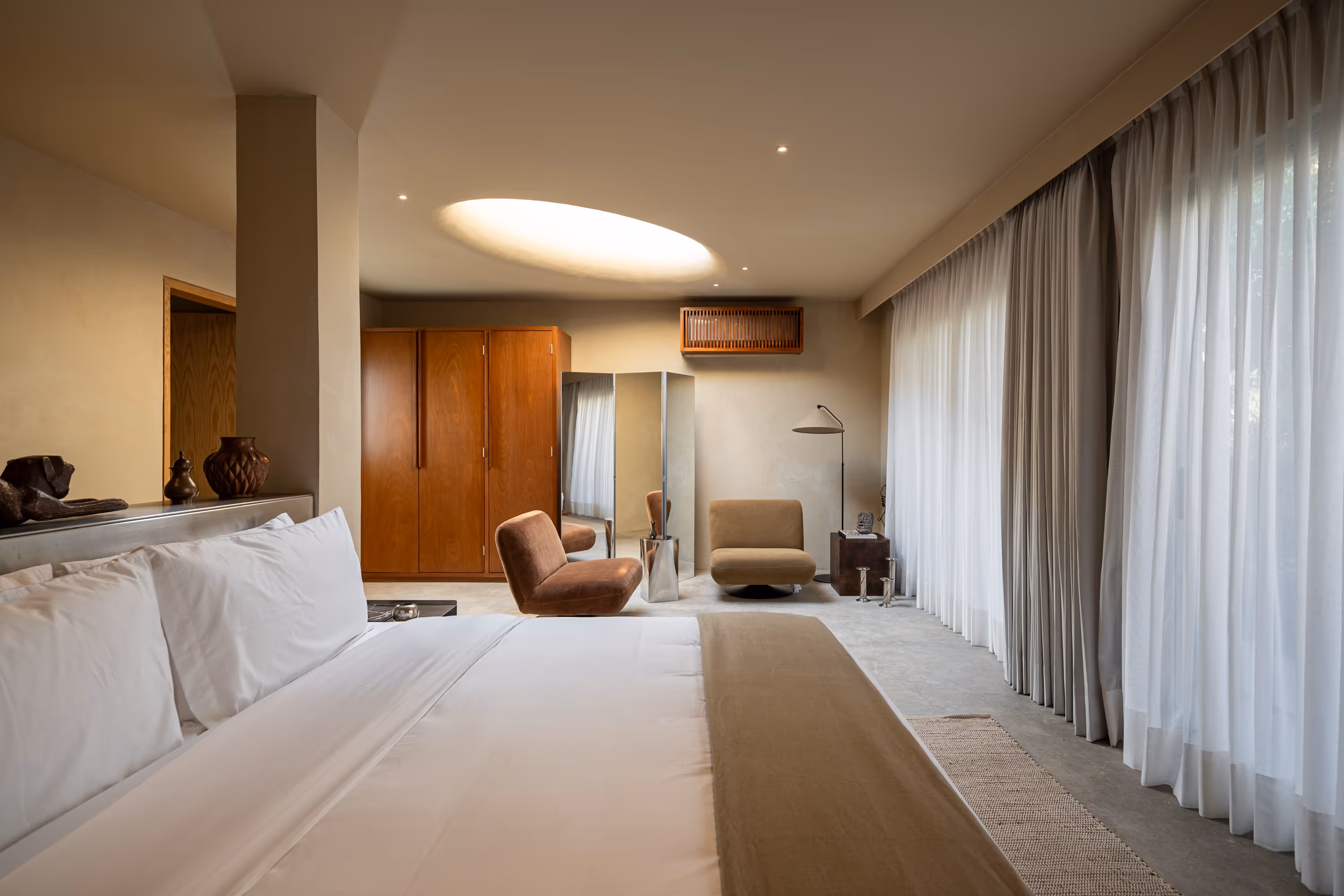 Modern bedroom with large bed, two upholstered chairs, wooden wardrobe, floor lamp, and sheer curtains on a wide window.