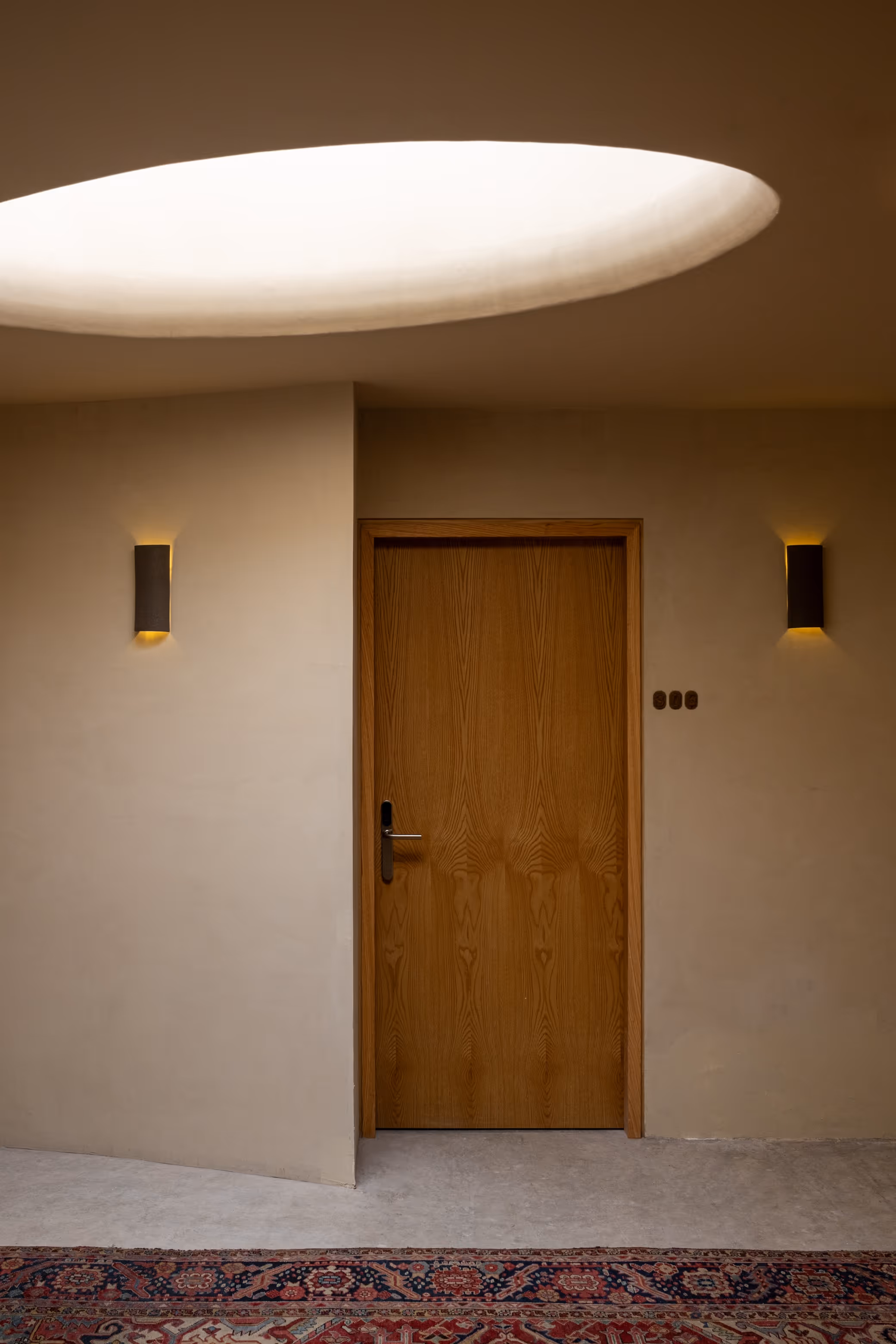 Wooden door framed by beige walls with two small wall sconces and an oval skylight above, with a patterned rug on the floor.