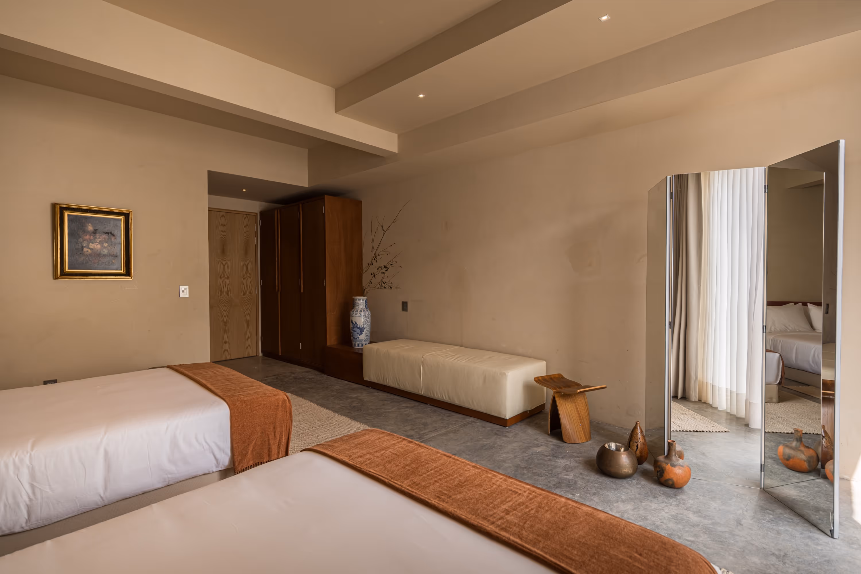 Minimalist bedroom with two beds featuring white linens and brown throws, a wooden wardrobe, a large wall mirror, and decorative vases on a polished concrete floor.