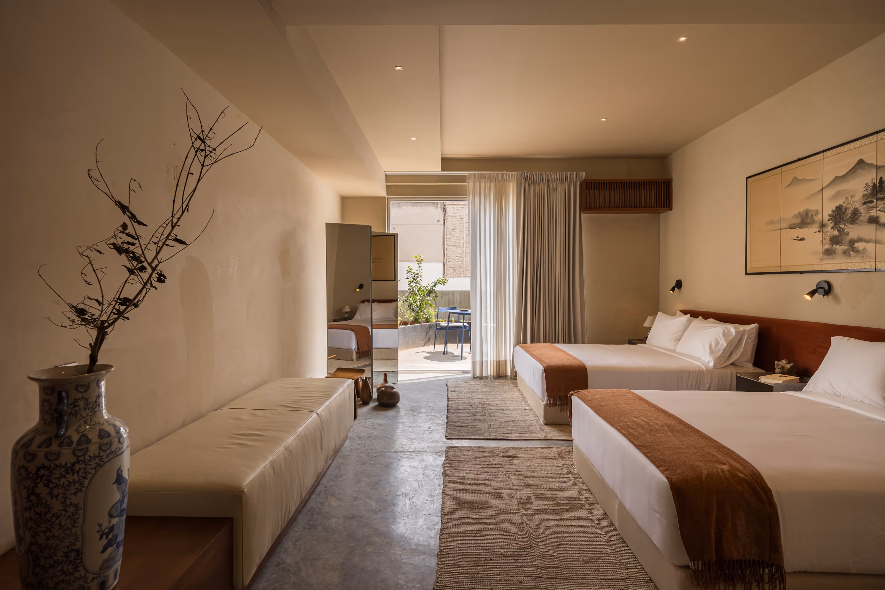 Hotel room with two double beds, brown throws, beige bench, and glass door leading to a small patio with table and chairs.
