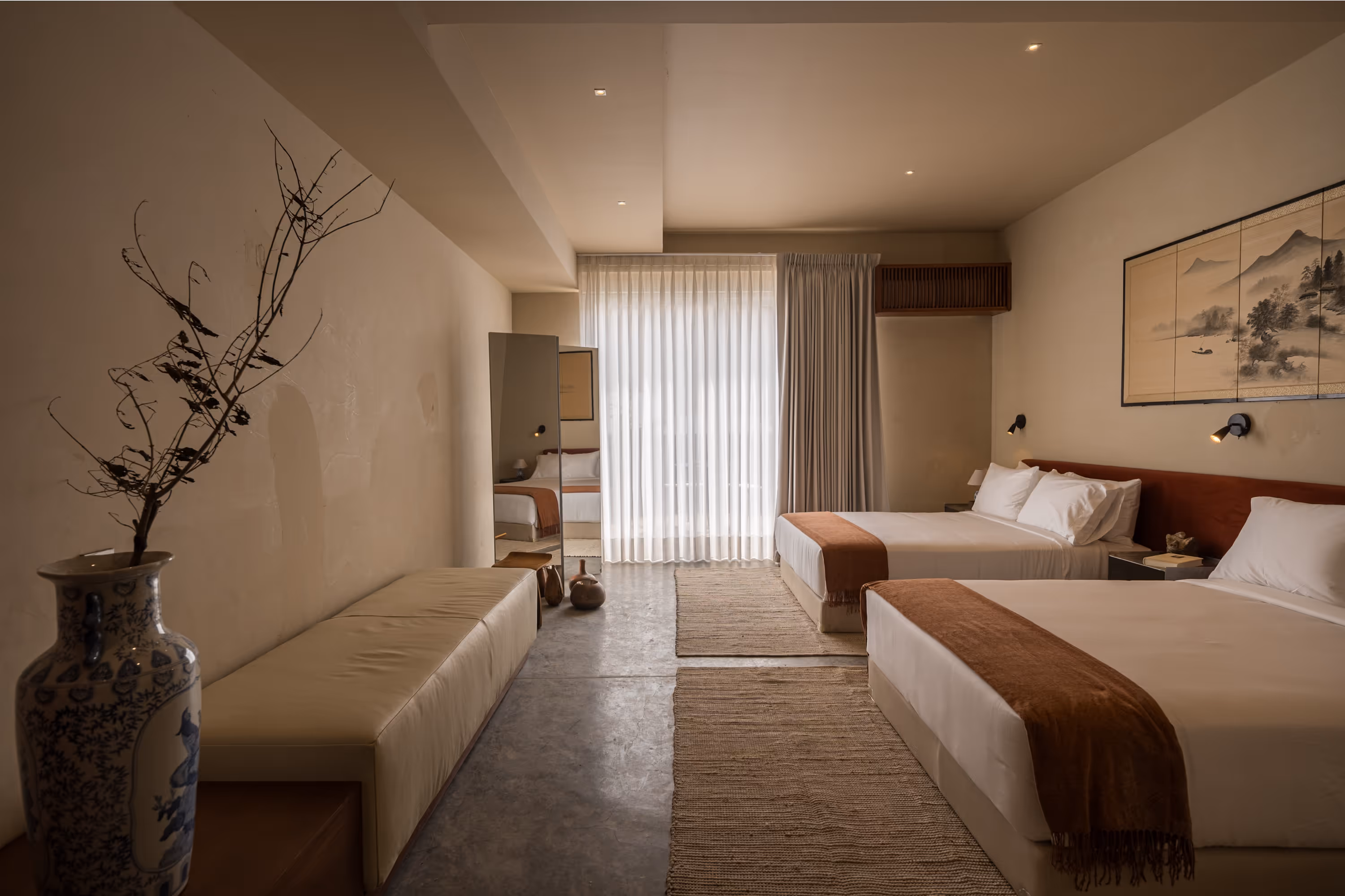 Minimalist hotel room with two double beds, beige walls, brown throws, a long bench, and a large vase with dried branches.