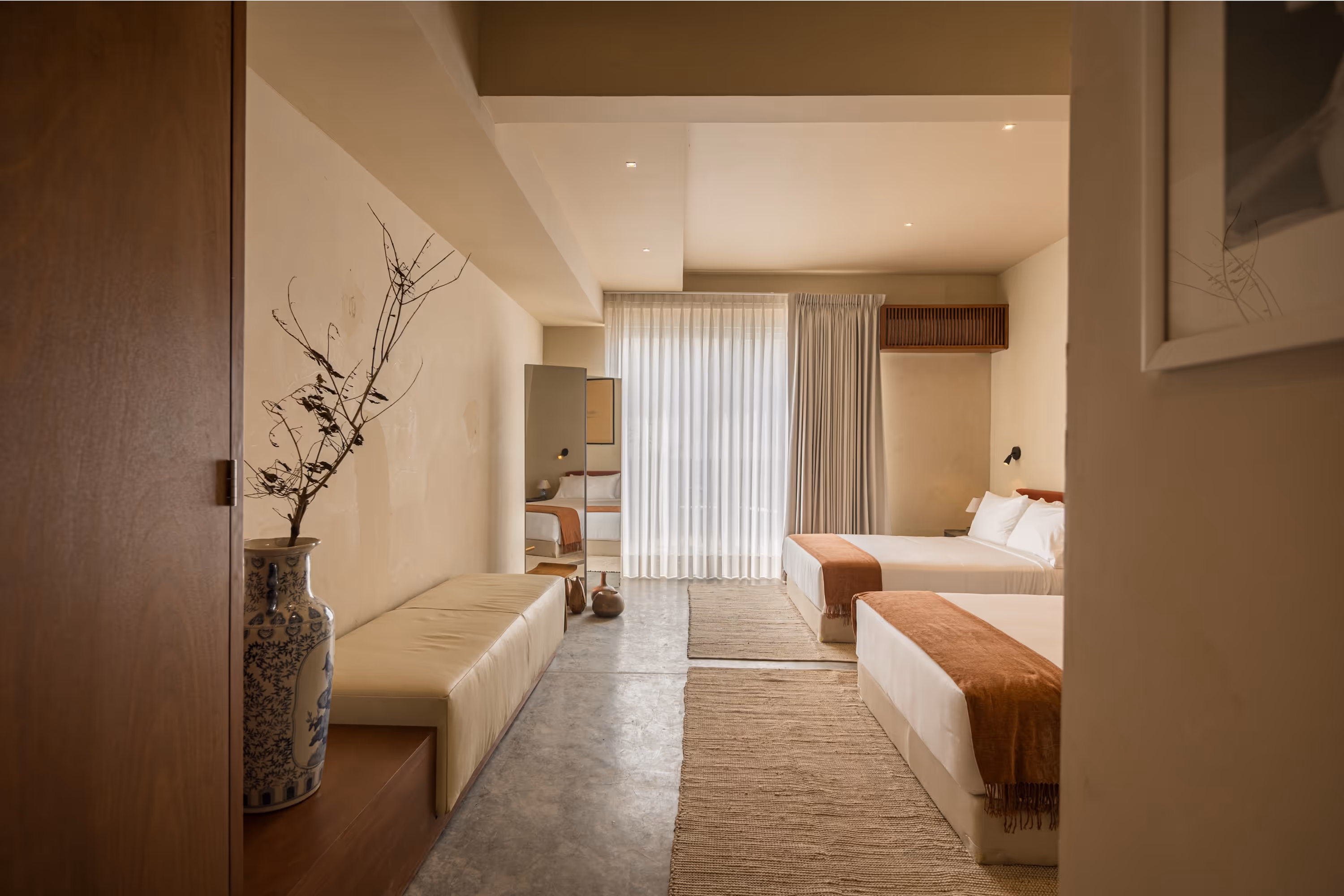 Minimalist hotel room with two double beds, beige walls, sheer curtains, and a decorative vase with branches.