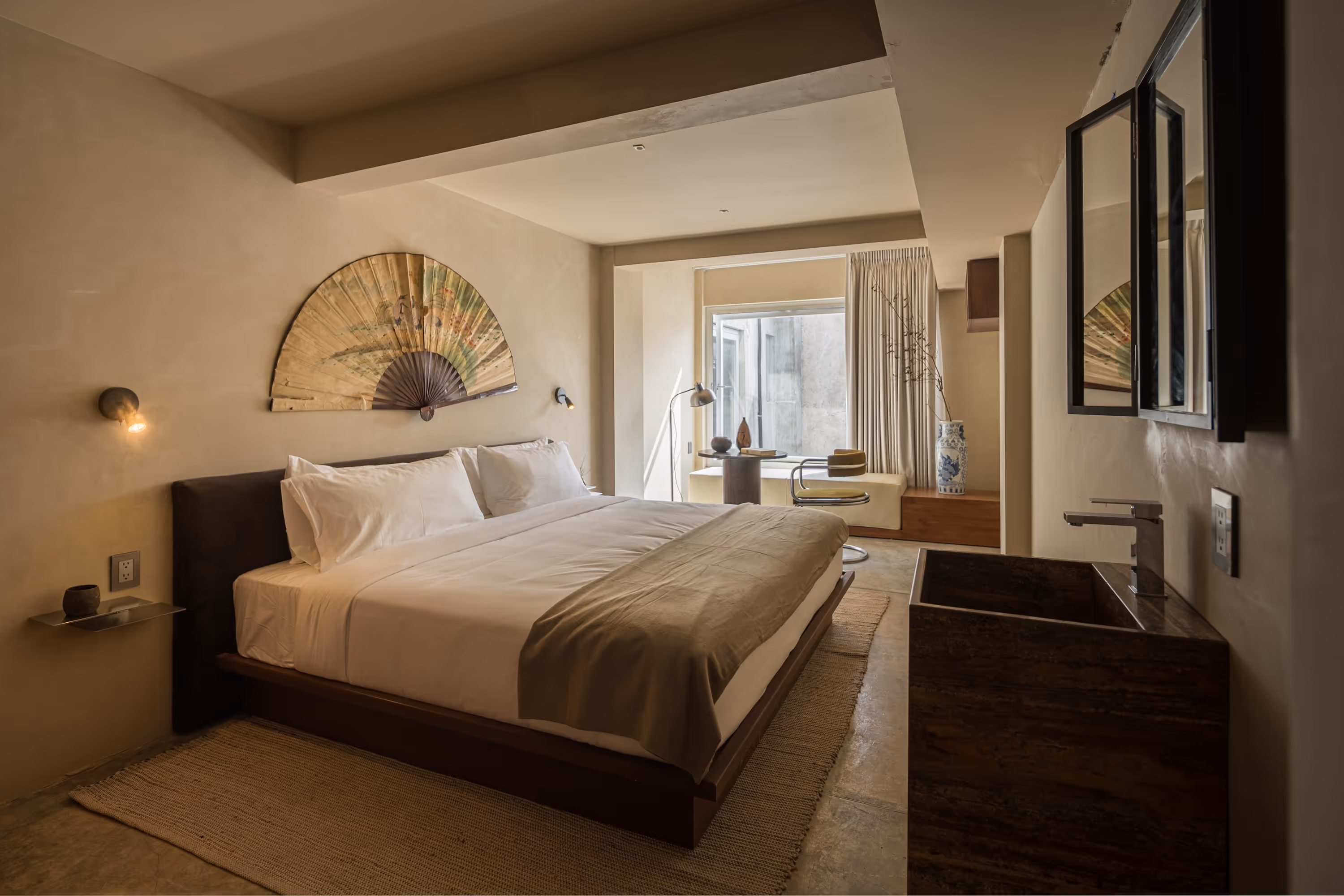 Minimalist bedroom with a large bed, beige bedding, wall-mounted fan decoration, a floor lamp, and a window seating area with a chair and ceramic vase.