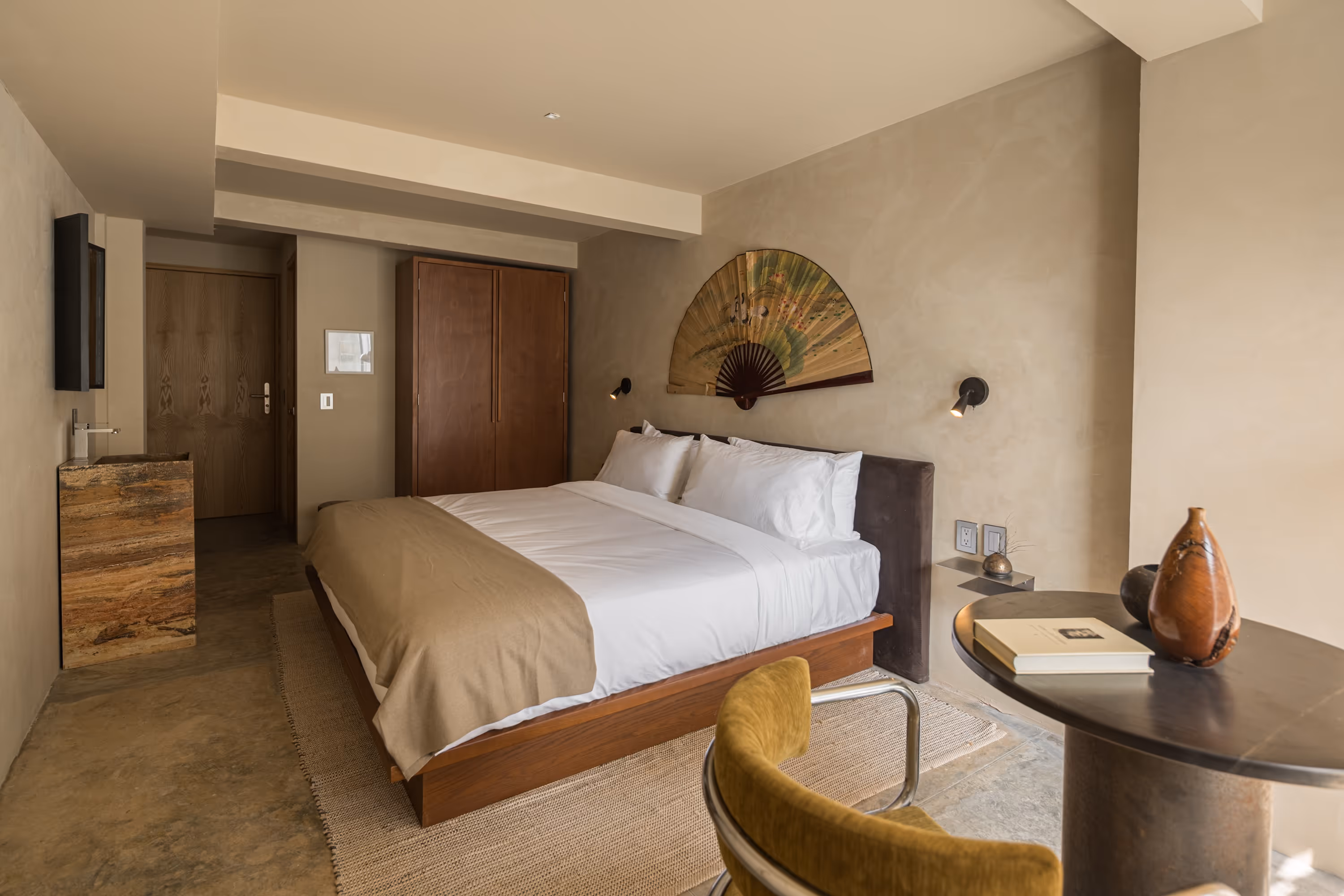 Minimalist bedroom with a double bed, beige blanket, wooden wardrobe, wall-mounted decorative fan, and round table with book and vases.