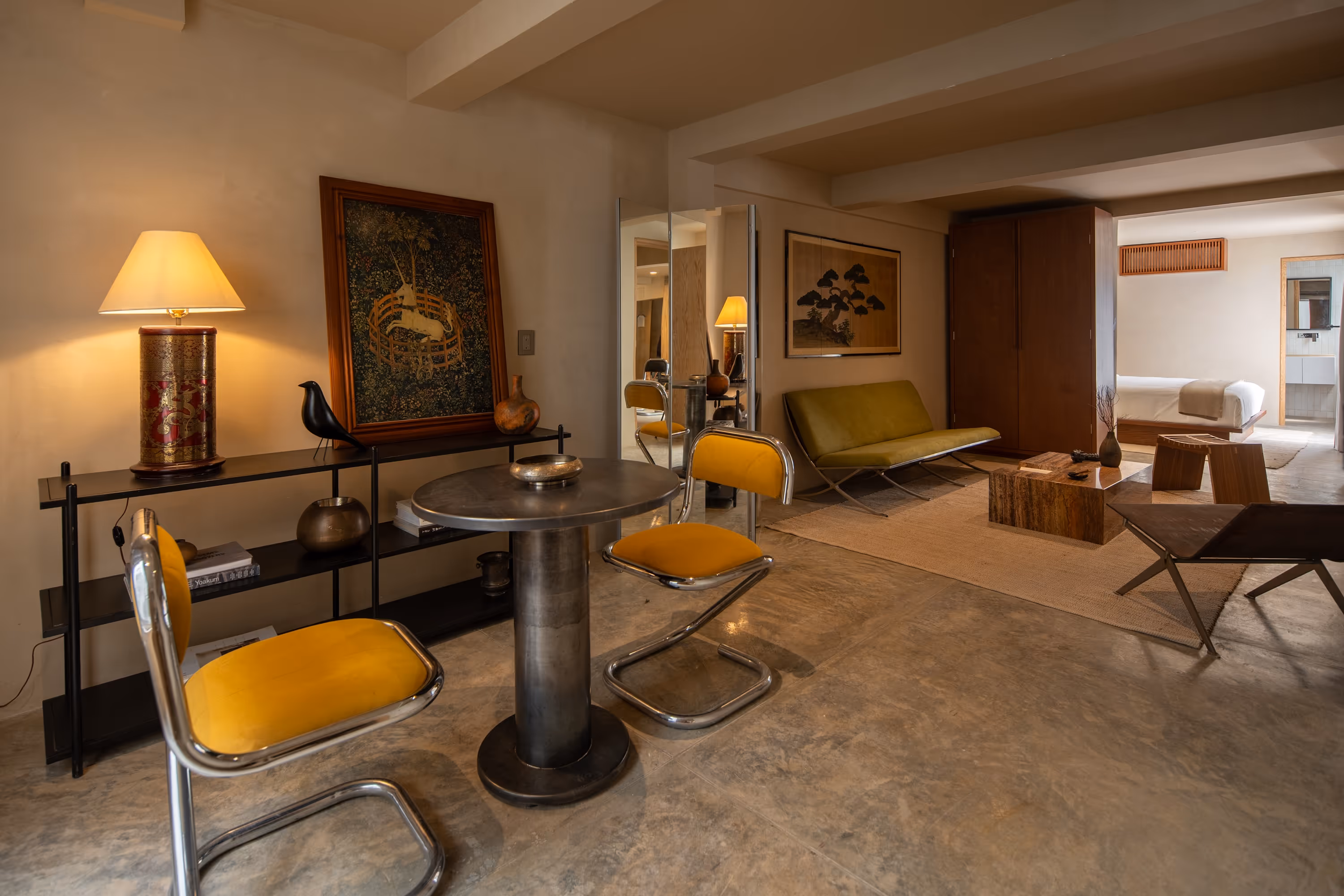 Modern living space with a round metal table, two yellow cushioned chairs, a green sofa, decorative artwork, and a visible bedroom in the background.