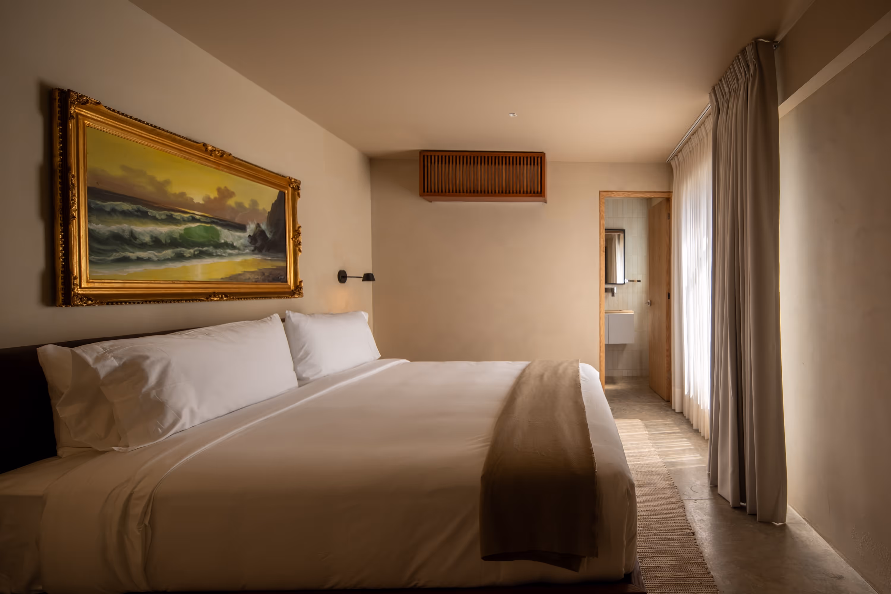 Minimalist bedroom with a king-sized bed, white linens, a brown throw, a framed seascape painting, and a view into a bathroom.
