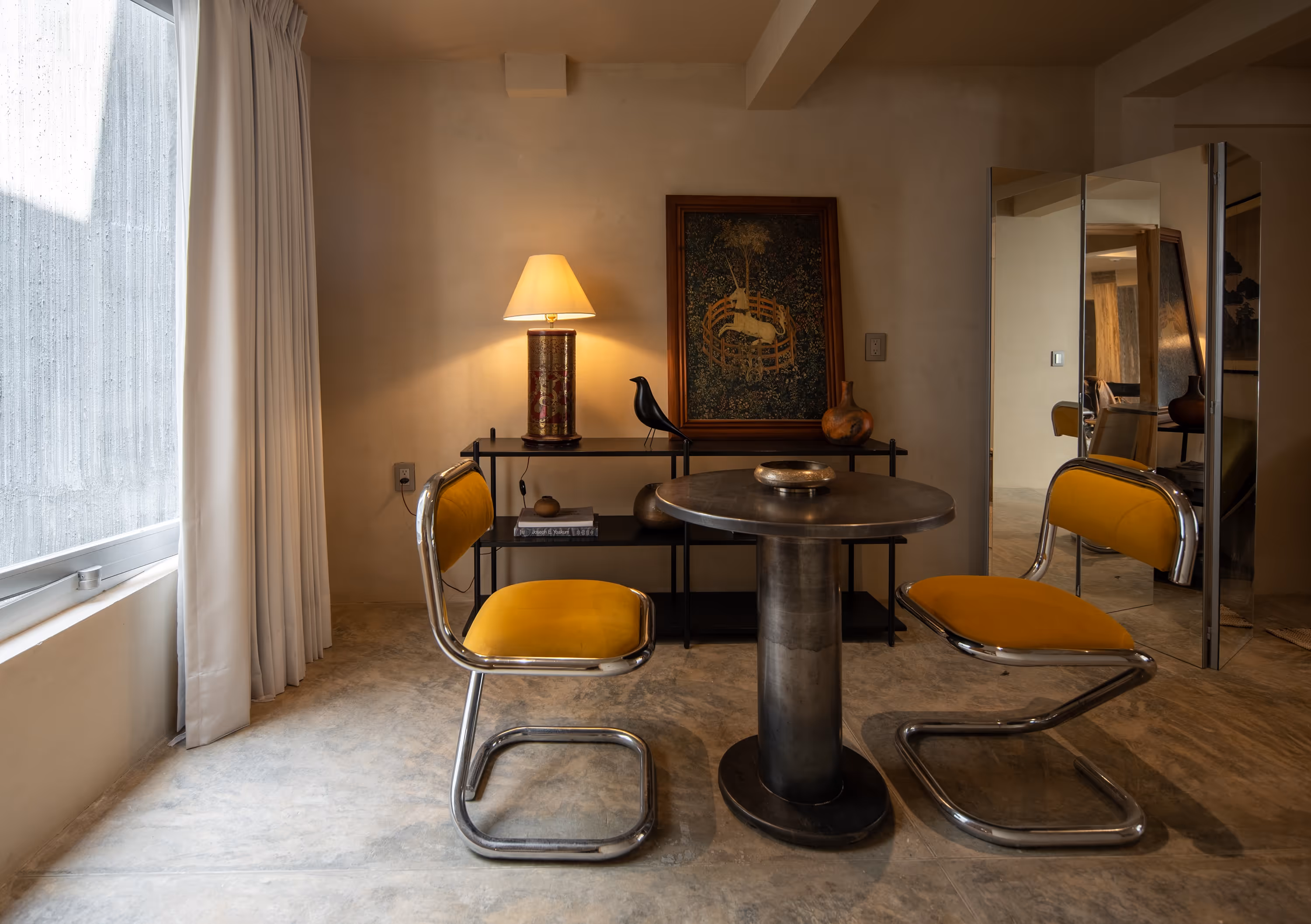 Cozy corner with round metal table, two yellow cushioned chairs, a lamp on a black shelf, and framed artwork on the wall.