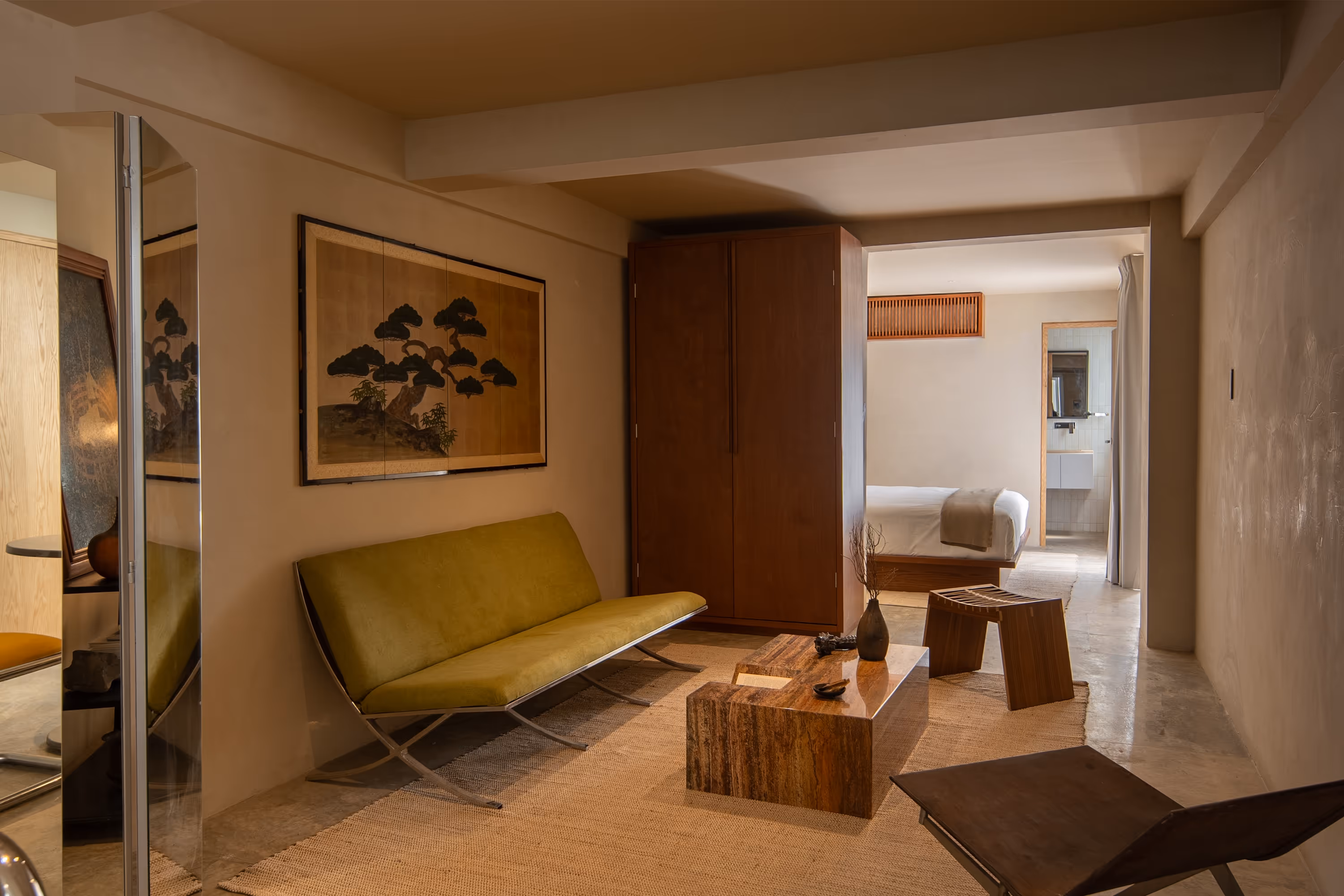 Minimalist hotel suite with green couch, wooden coffee table, wooden stool, and view into bedroom with white bed and bathroom.