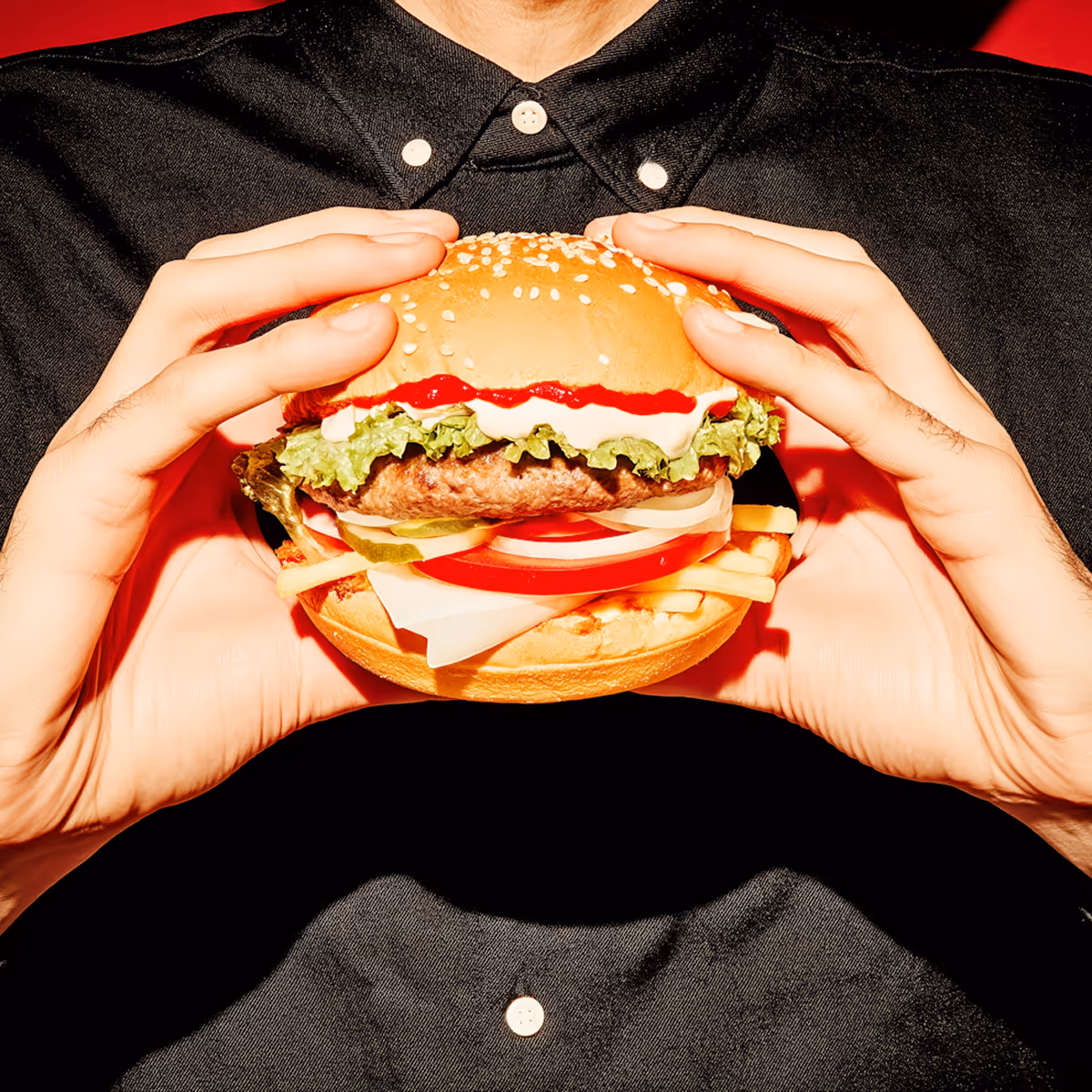 Person in black shirt holding a hamburger with lettuce, cheese, tomatoes, pickles, onions, ketchup, and mayonnaise.