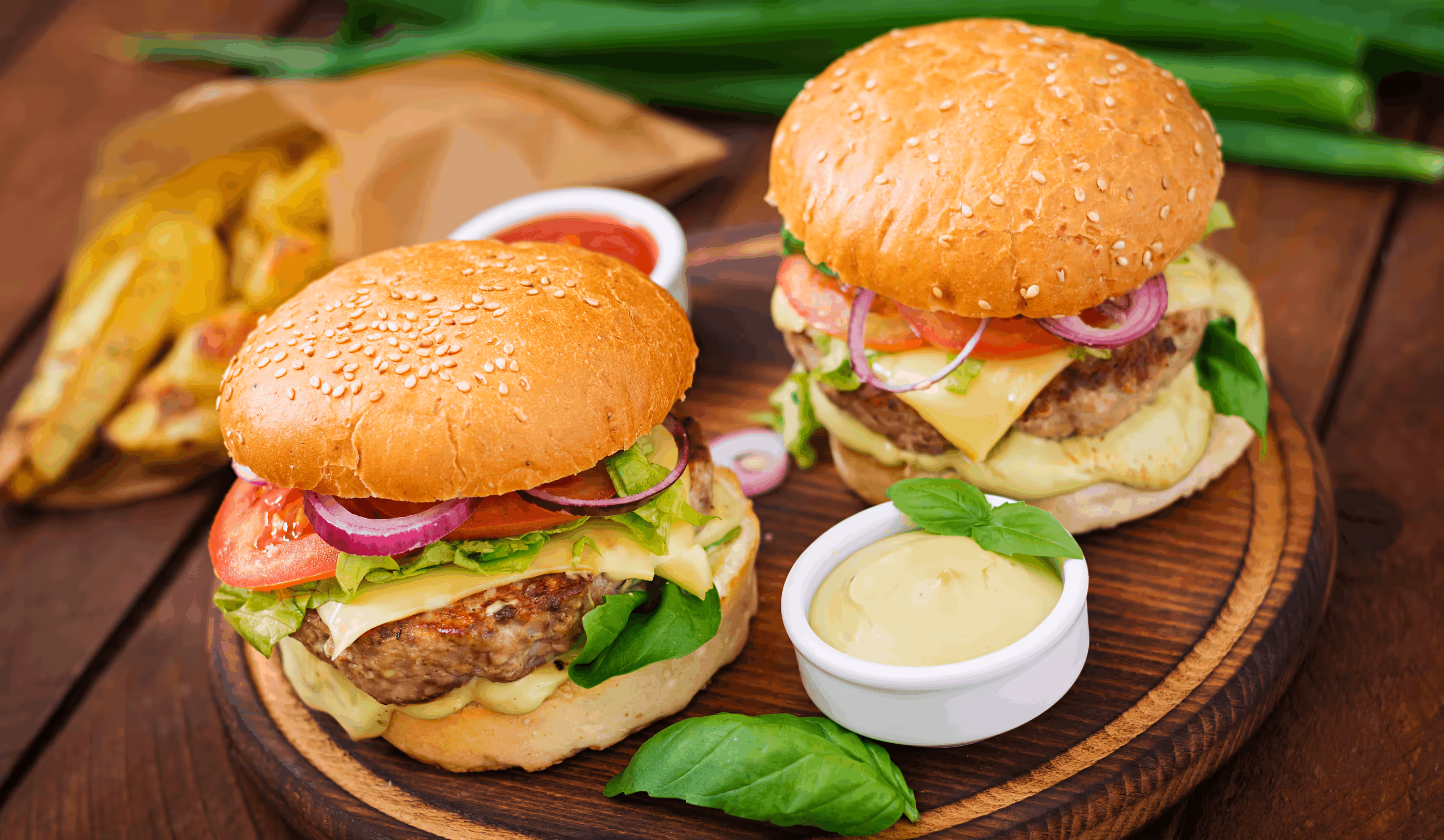 Two cheeseburgers with lettuce, tomato, red onion, and sauce on sesame buns served on a wooden board with dipping sauces and fresh basil leaves.