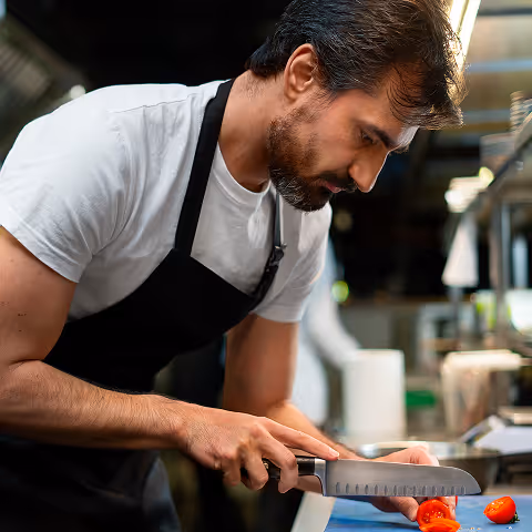 Man in a black apron and white shirt slicing a red tomato with a knife in a kitchen.
