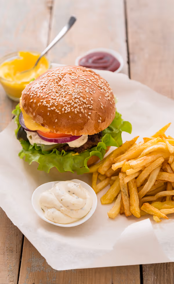 Cheeseburger with lettuce, tomato, onion, and sauce served with golden French fries and dipping sauces on a wooden table.