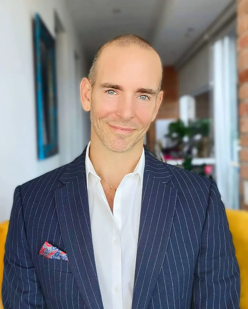 Smiling bald man with blue eyes wearing a navy pinstripe suit jacket and white shirt indoors.