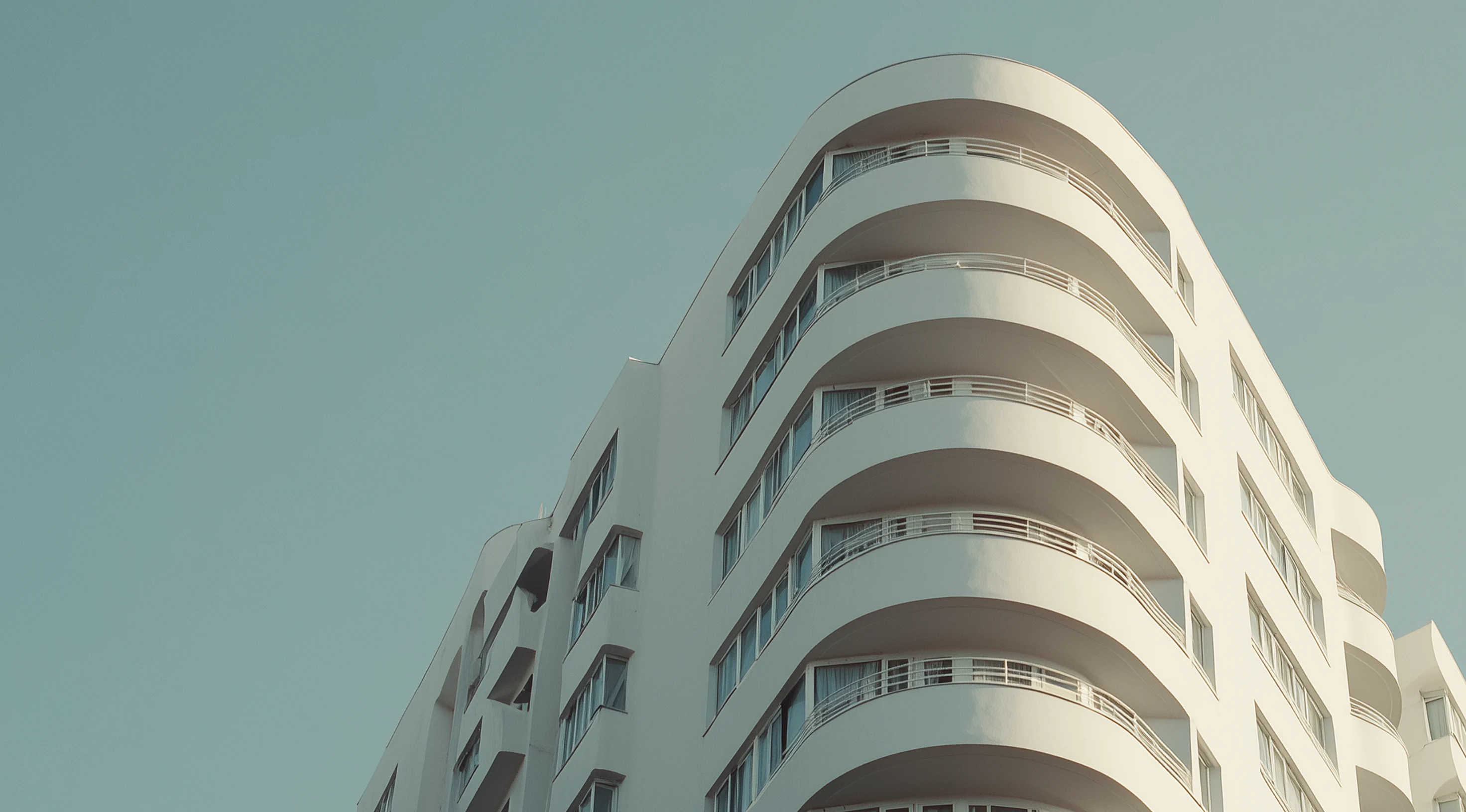 A modern white building with curved balconies rises against a clear blue sky, showcasing sleek architecture and stylish design.