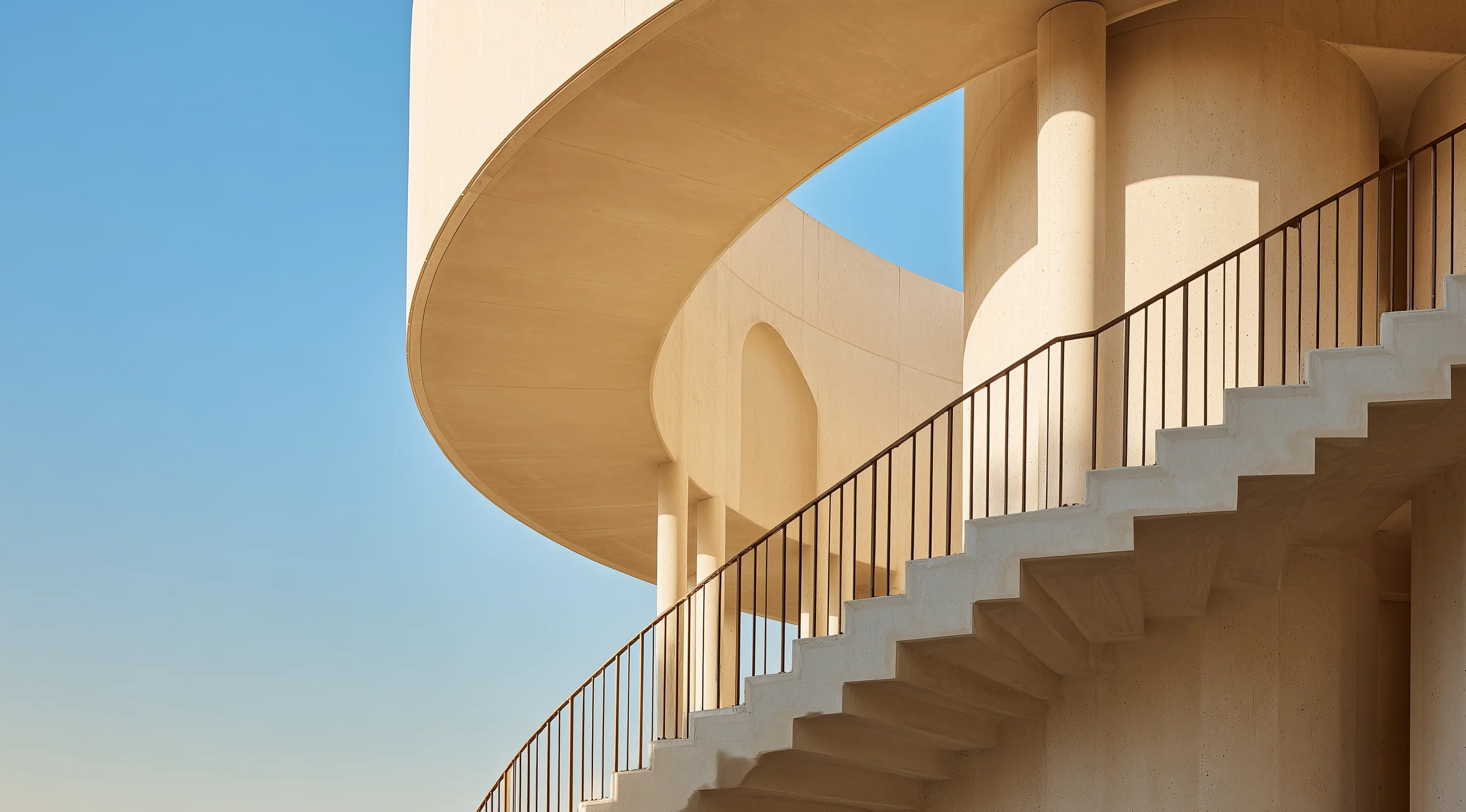 A close-up of a modern architectural staircase with curved railings and beige concrete, against a clear blue sky.