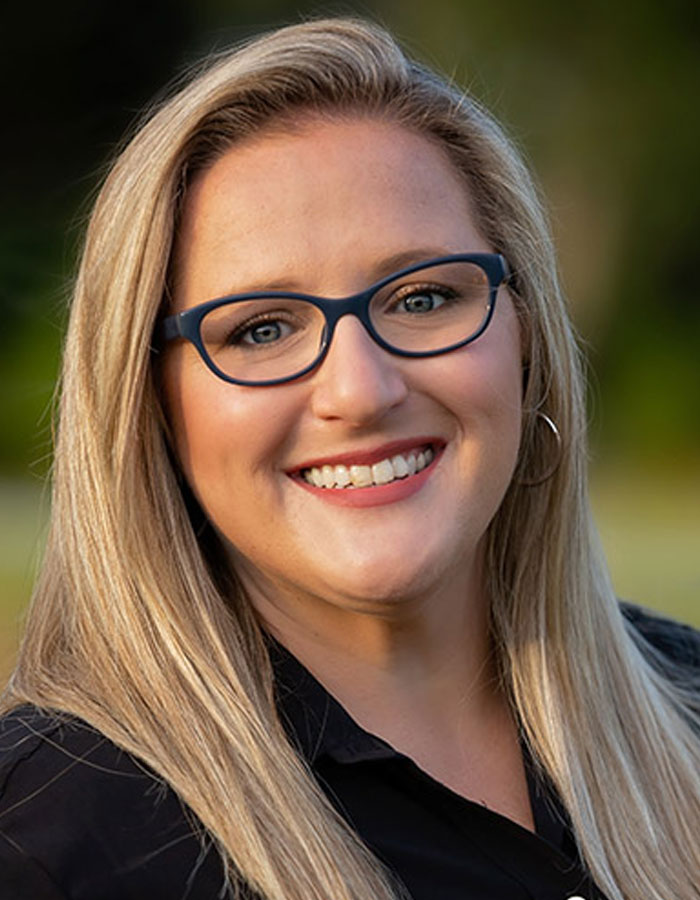 Kristin Macari with long blonde hair, blue rectangular glasses, and hoop earrings, wearing a black top outdoors.