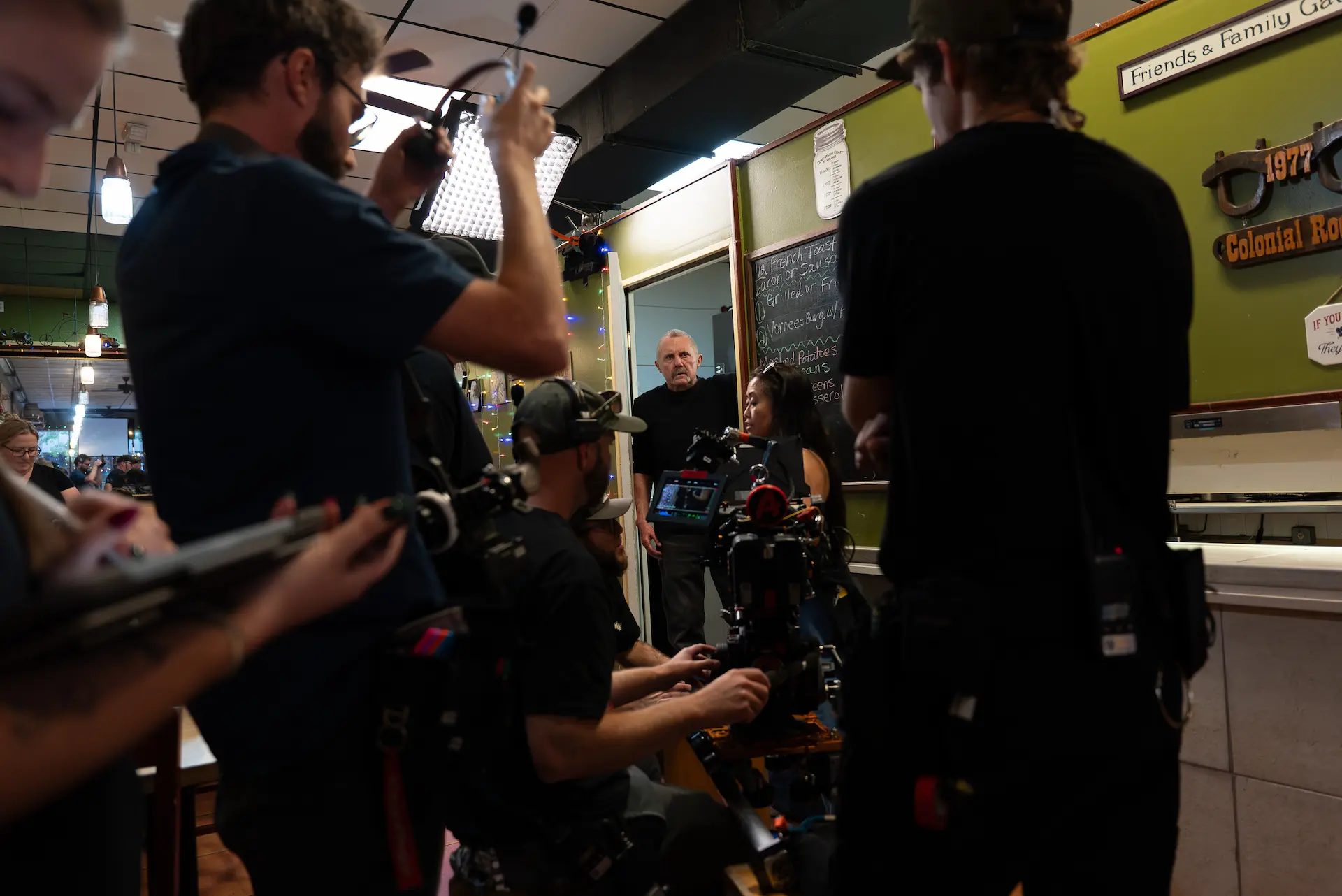 Film crew setting up a scene in a restaurant with a man standing in a doorway and a woman sitting inside.