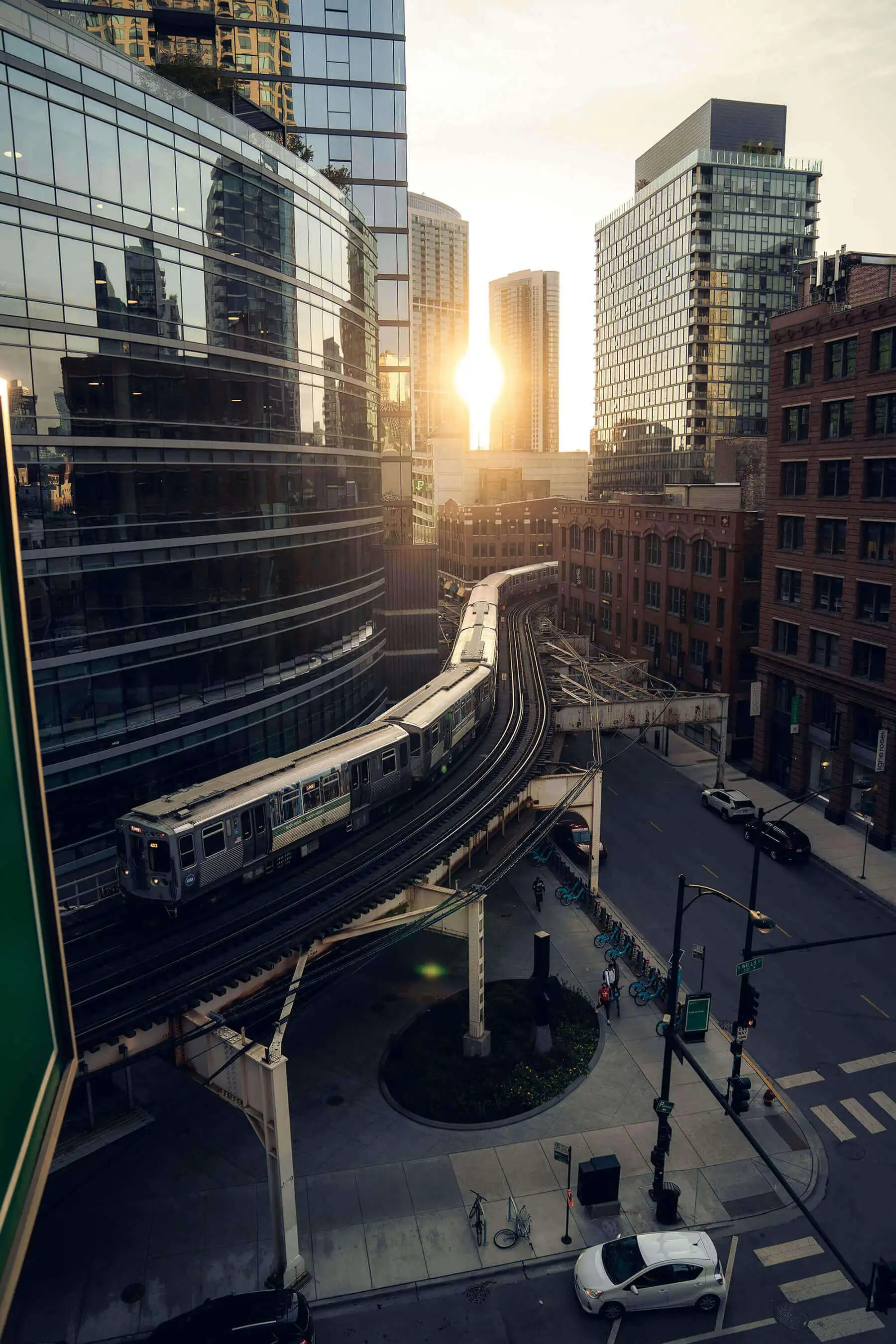 Elevated train curving through downtown city streets at sunset with modern glass and older brick buildings.