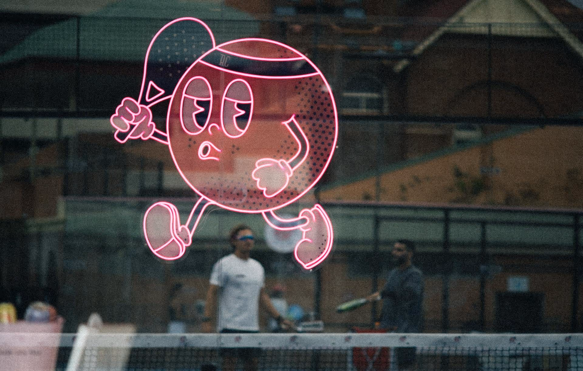 Pink neon sign of a cartoon tennis ball holding a racket, with two men playing tennis behind a glass partition.