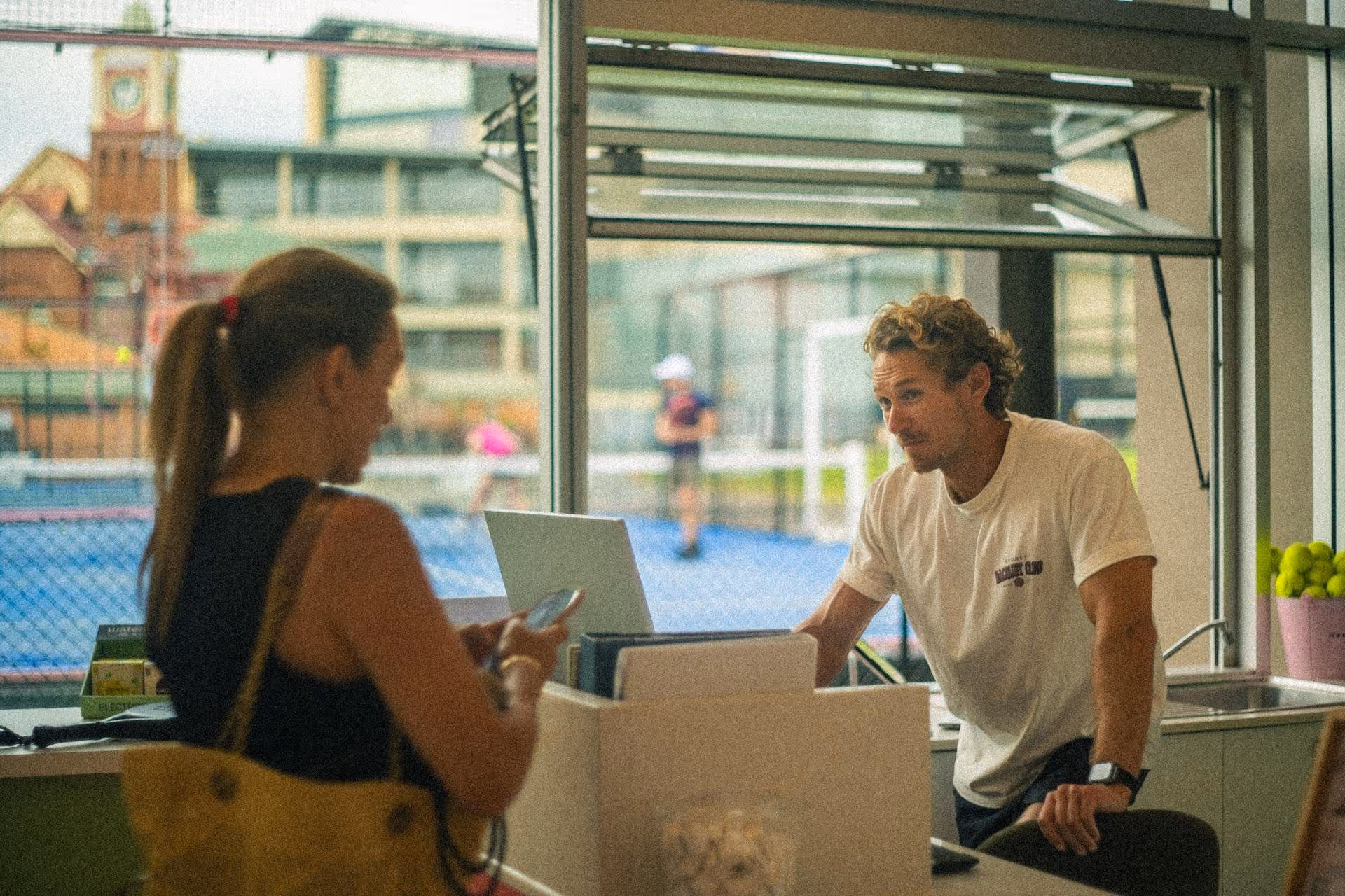 Man in a white t-shirt leaning on a counter talking to a woman with a ponytail holding a phone inside a sports facility with a tennis court visible through the window.