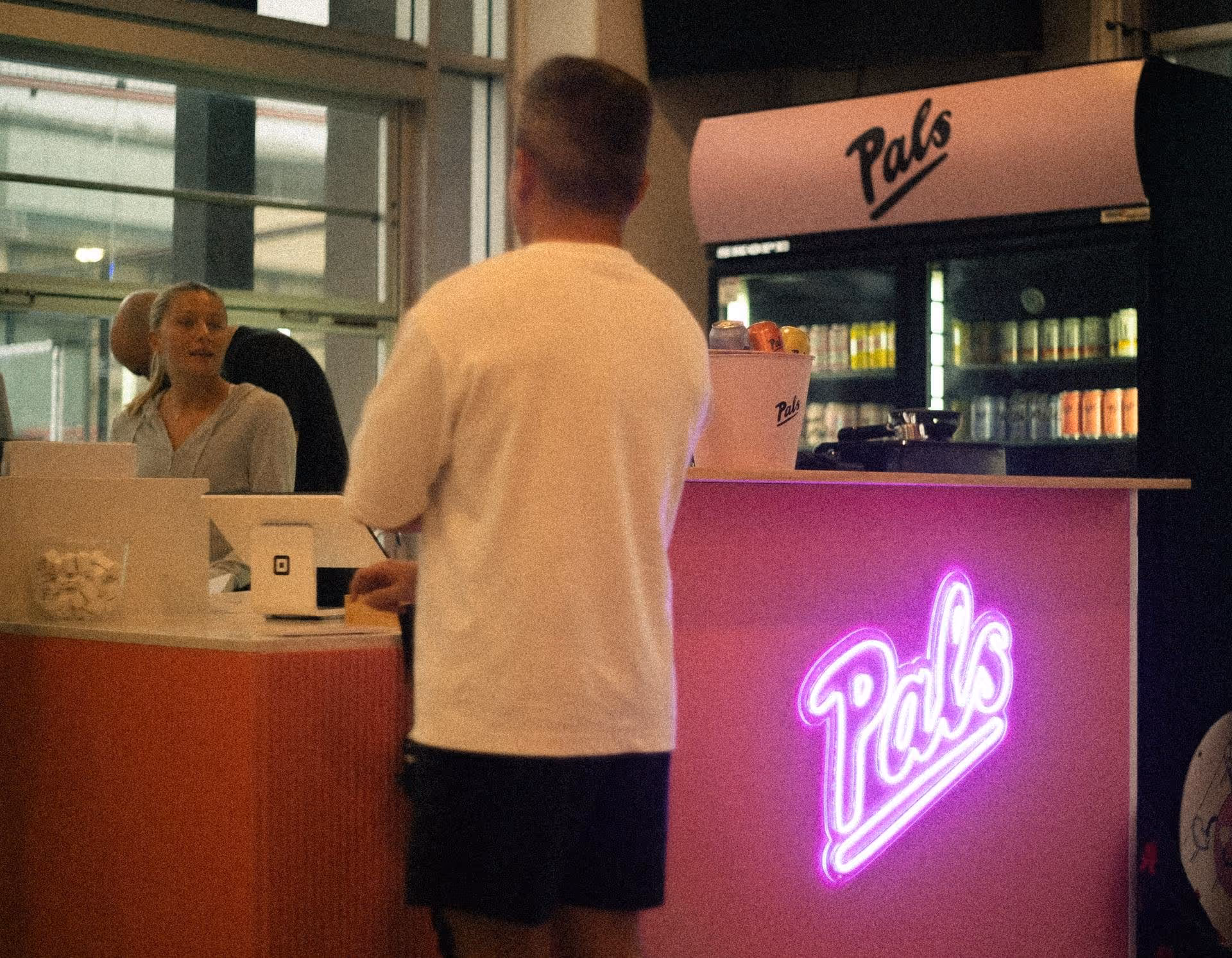 Man in white shirt ordering at a pink Pals counter with a female attendant and a refrigerator stocked with drinks in the background.