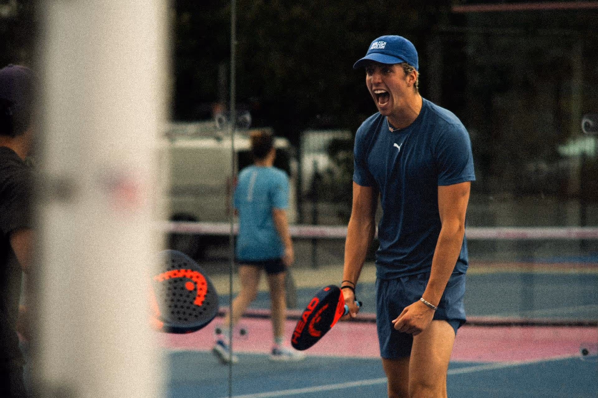Young man wearing a blue shirt and cap enthusiastically shouting while holding a padel racket on a padel court.