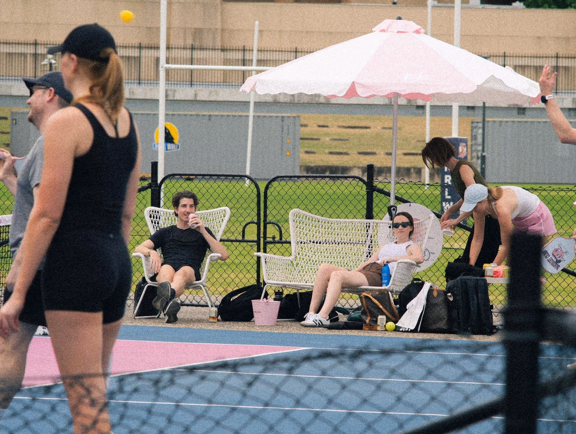 Group of people relaxing by a fenced pickleball court with a pink and white umbrella, some seated on white wicker chairs and others standing nearby.